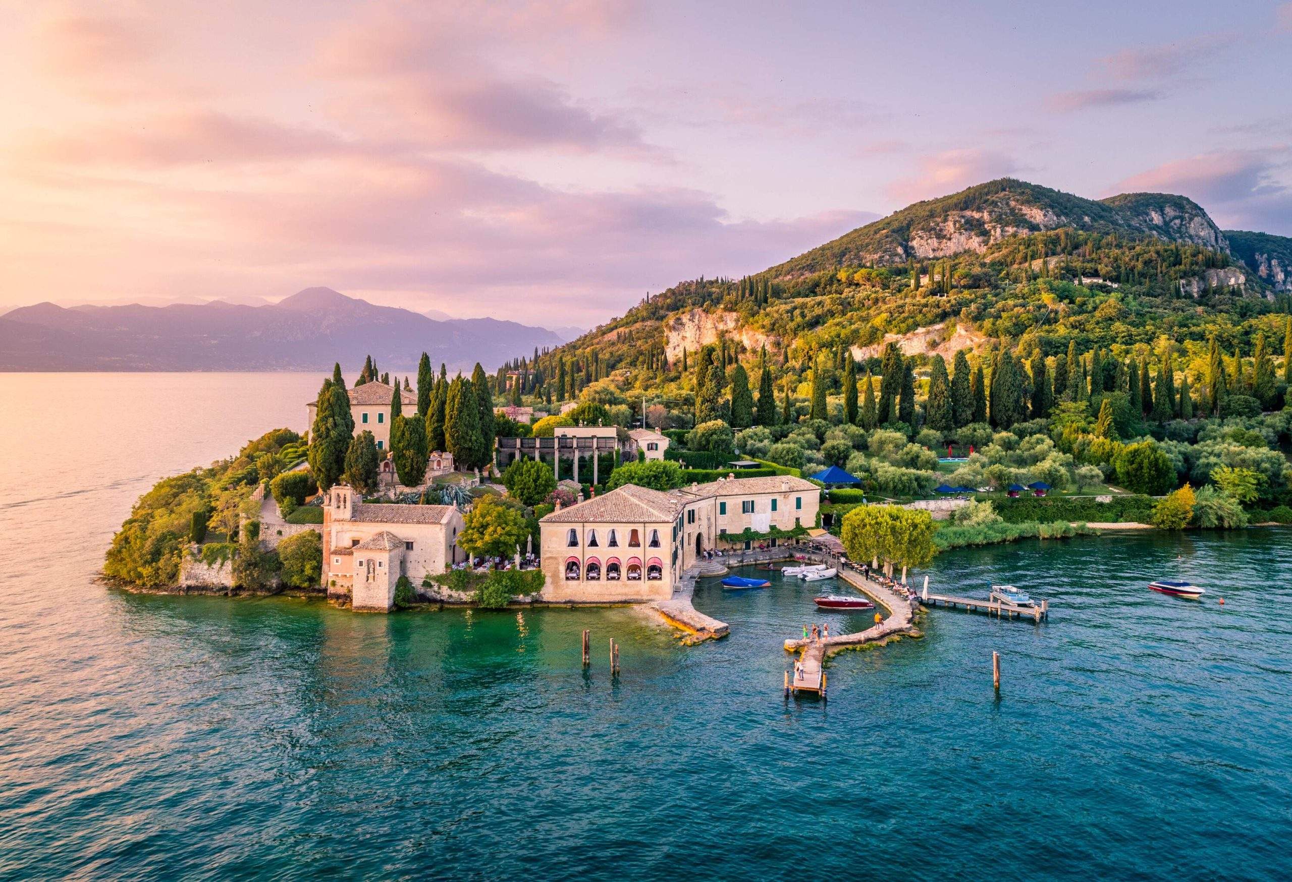 A classic resort building with a small harbour under a green forested hill against a quiet lake and purple sky.