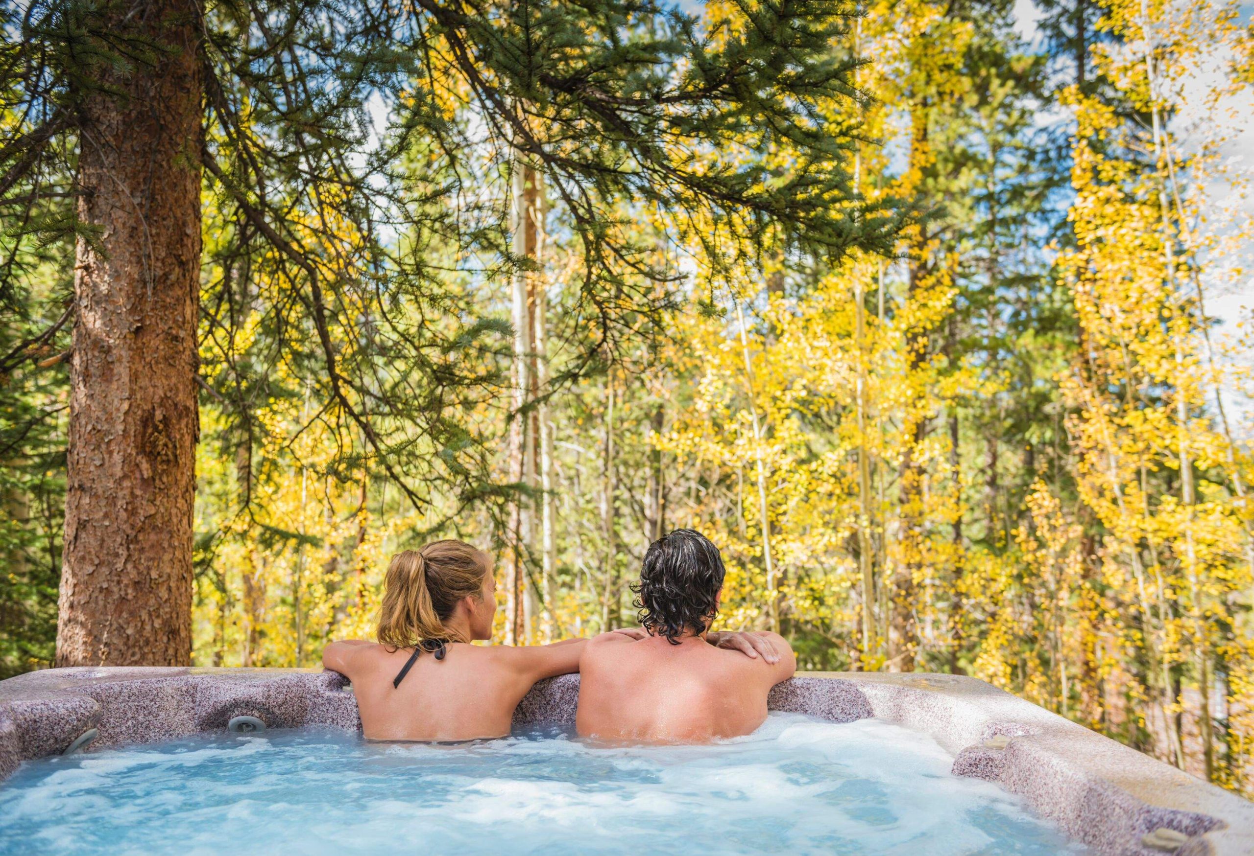 a man and a woman sitting in an outdoor hot tub looking out at the forrest