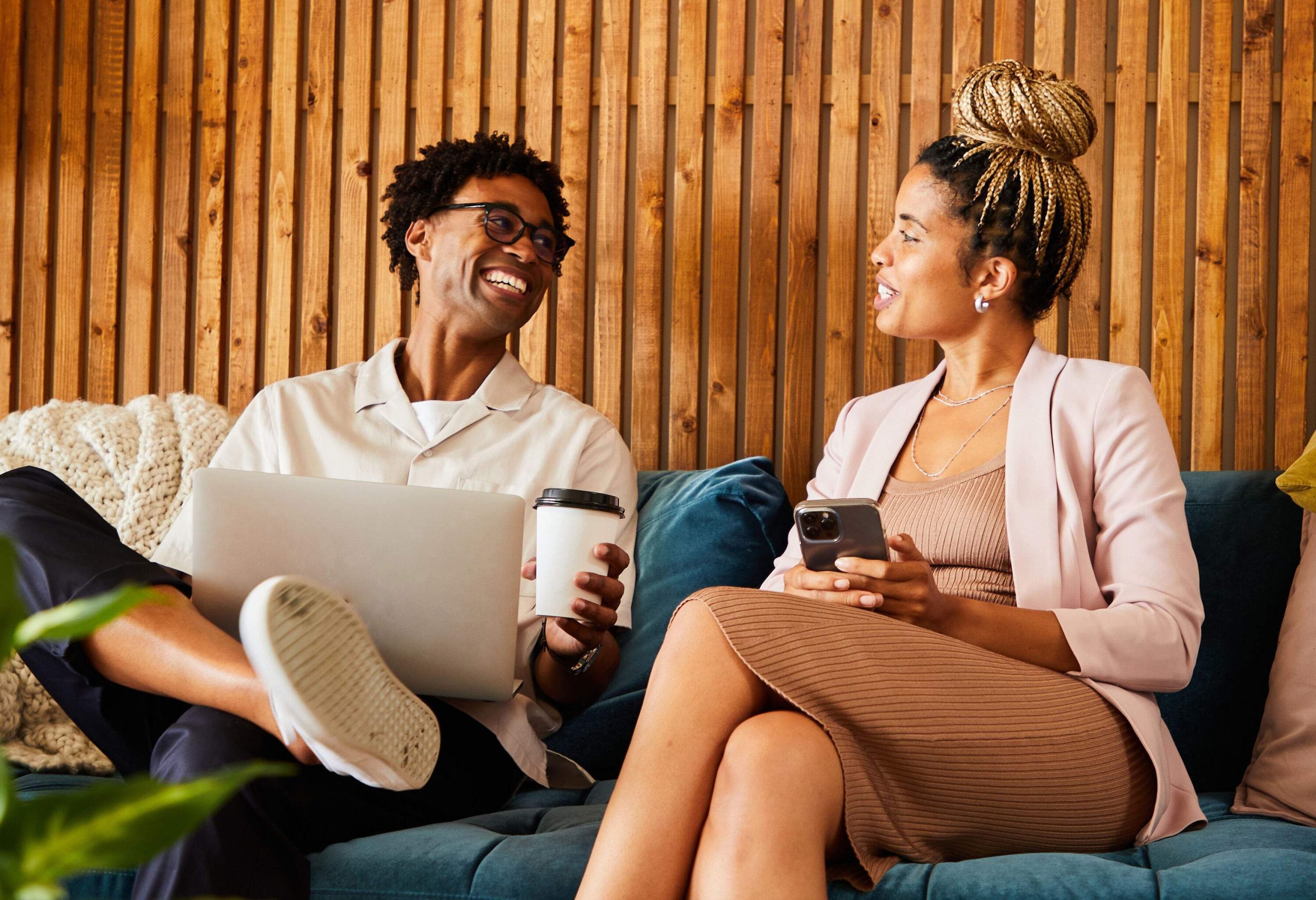 A woman holding a smartphone and a man with a coffee cup talking with each other while sitting on a blue couch.