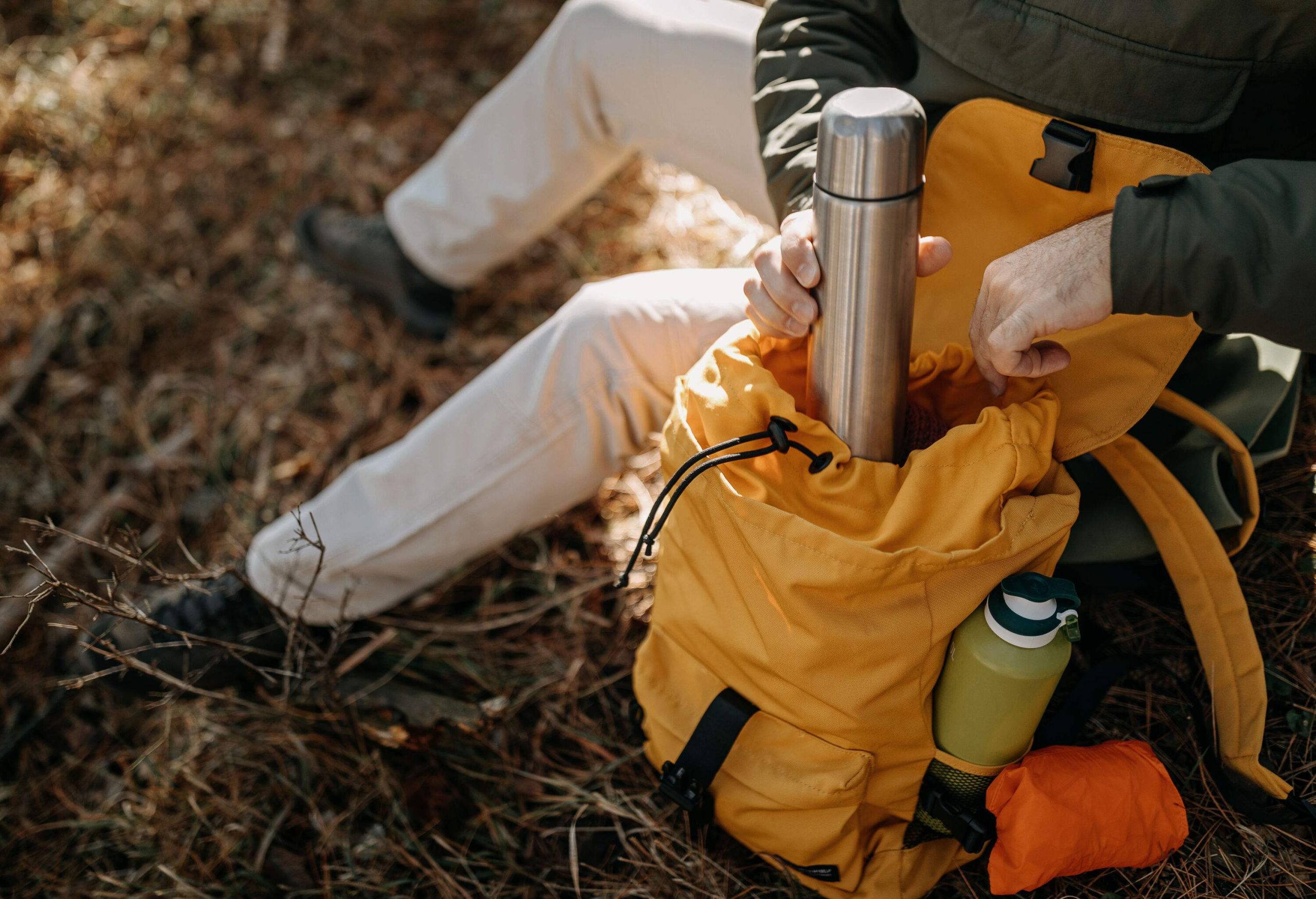 A man resting on the ground brings out a silver vacuum-insulated bottle from his yellow backpack.