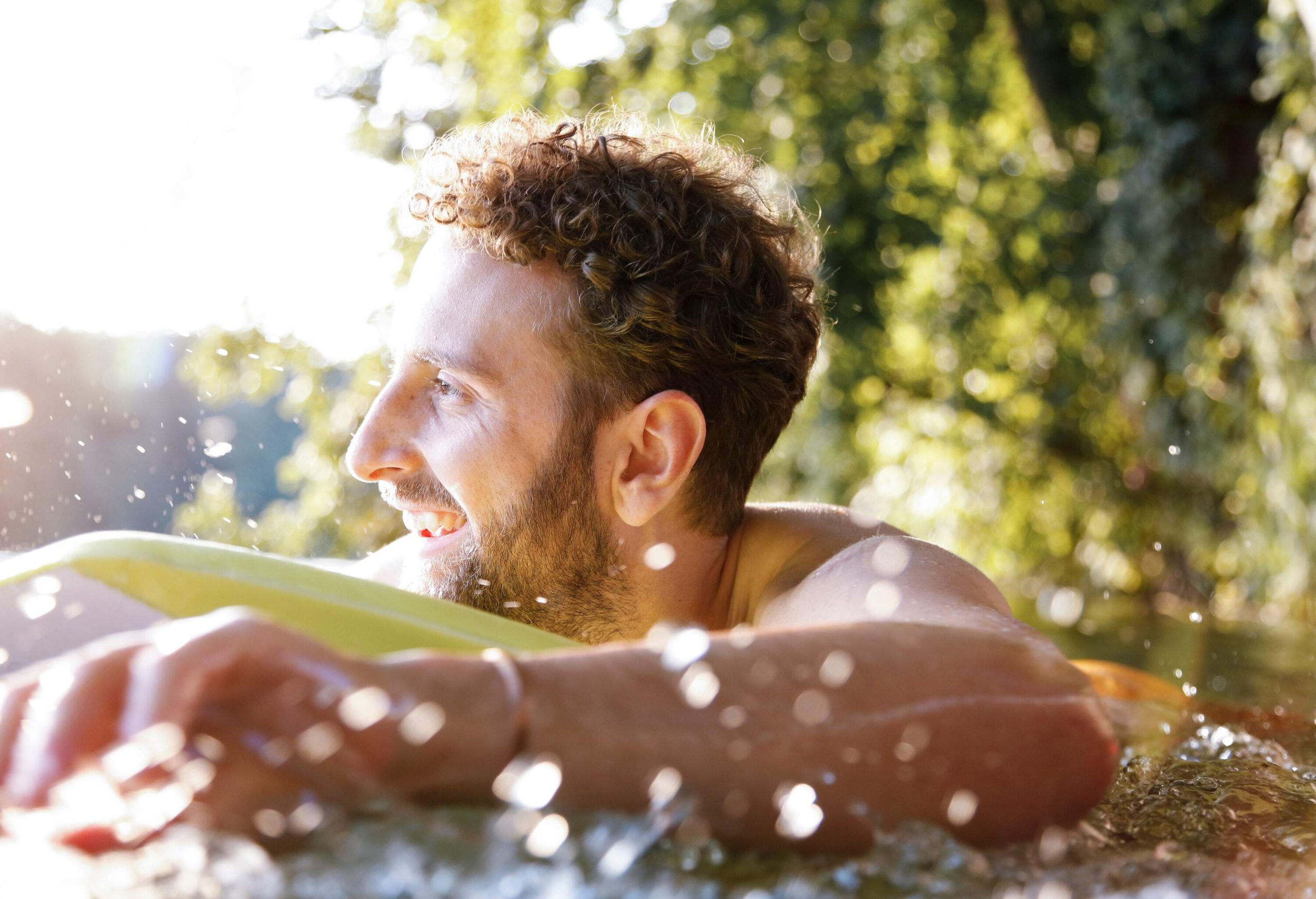 A bearded man smiles while hand paddling on a surfboard.