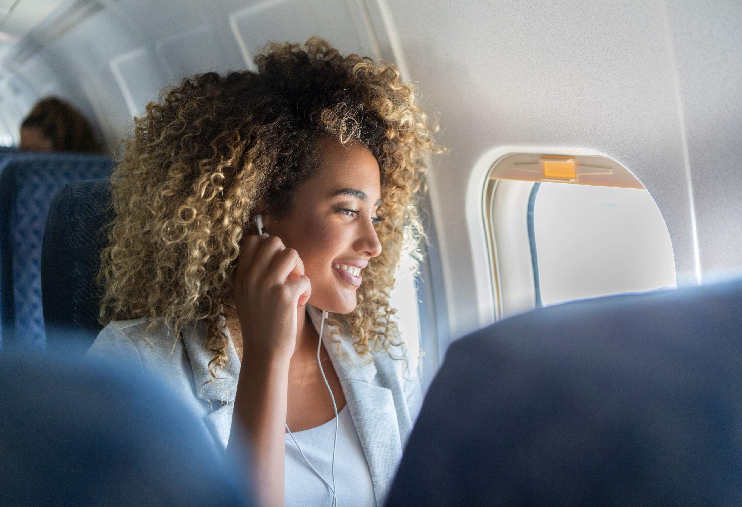 A woman with curly hair puts on her earbuds as she settles into her window seat on an airplane.