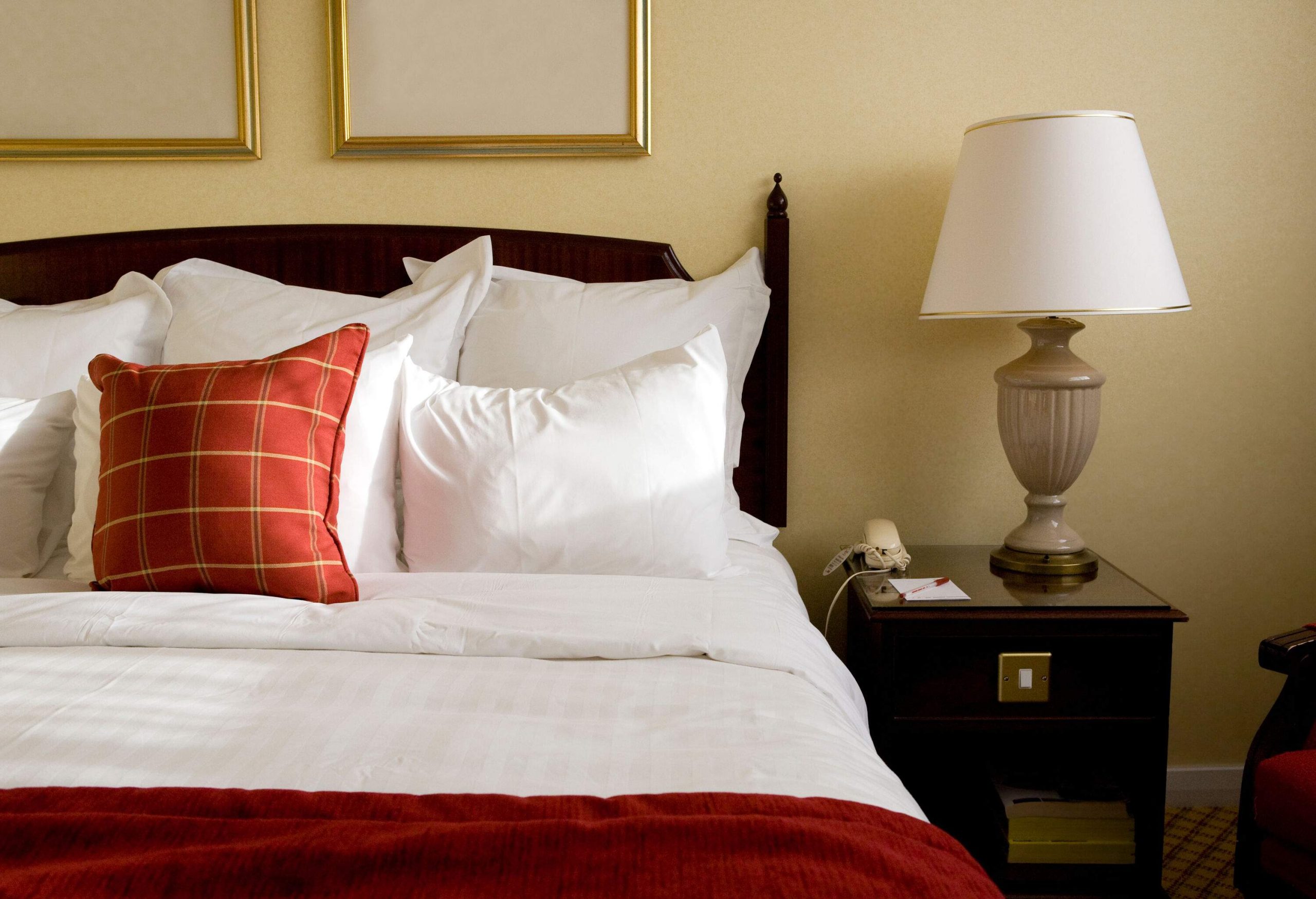 A bed with white linens and a red accent alongside a wooden table with a lamp and a wired phone