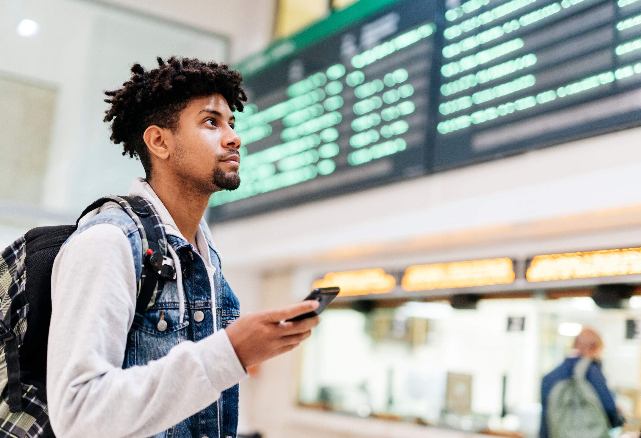 A young man holding hisA man wearing a denim vest and backpack stands in an airport, holding a mobile phone, and intently studying the departures board. smartphone checks the flight schedules on the airport's departure board.