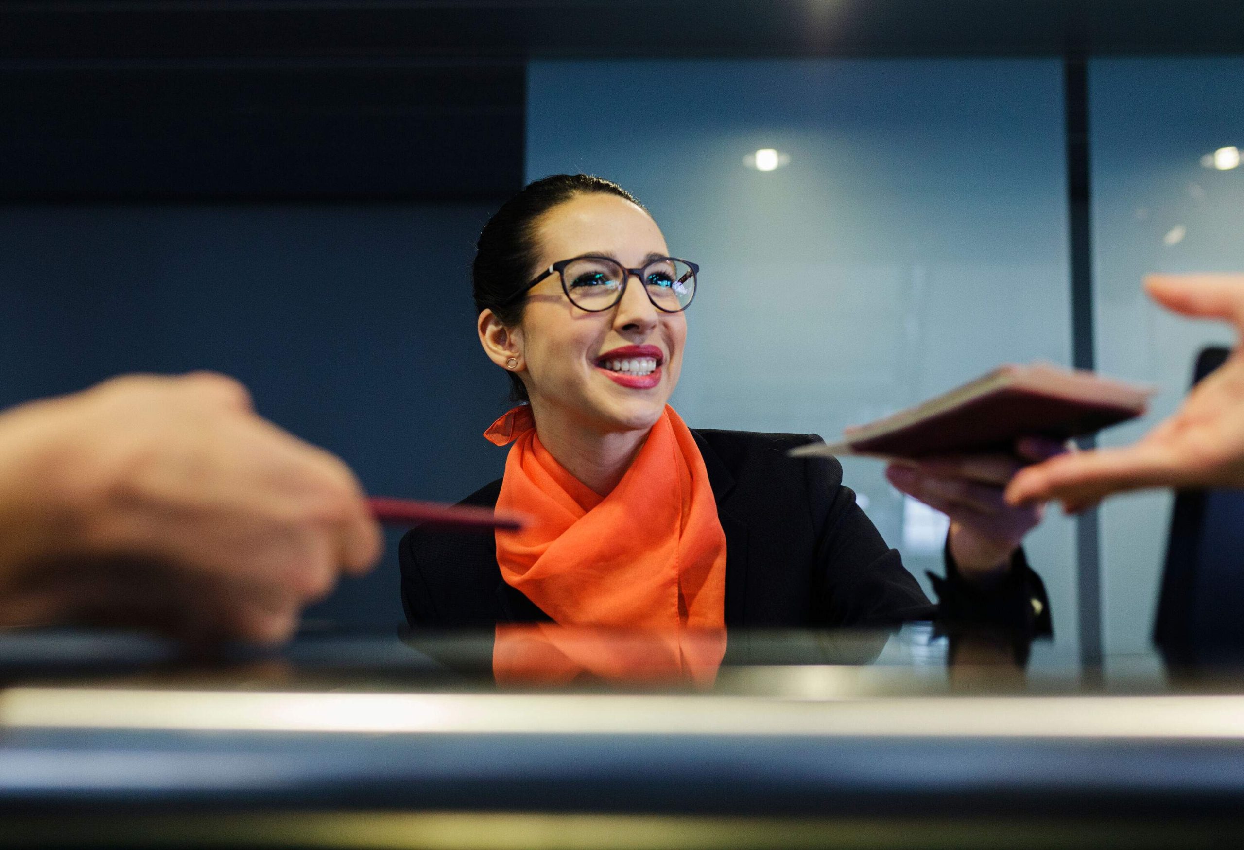 A lovely receptionist hands a passport to a traveller while one submits his at the counter.