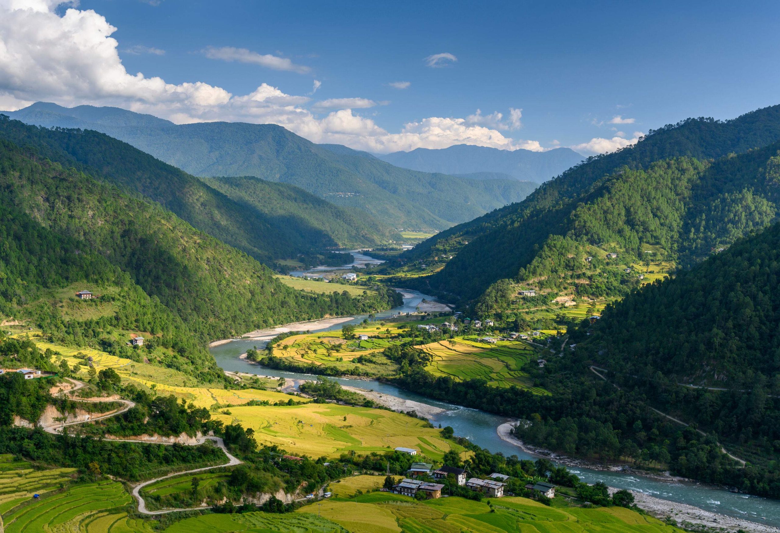 A meandering river gracefully snakes its way between densely wooded mountains, with thriving communities nestled at their feet and terraced farms cascading down the slopes.