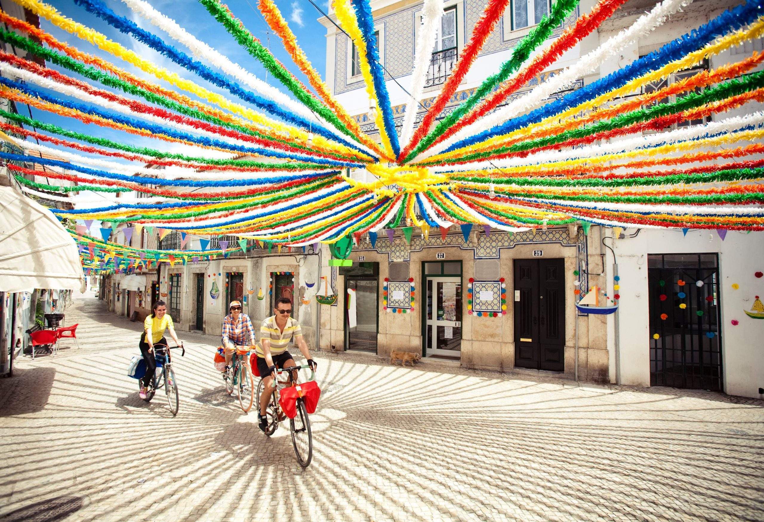 Three riders traverse a lively street where colourful festival streamers flutter overhead, adding to the festive atmosphere as a row of charming houses lines the bustling thoroughfare.