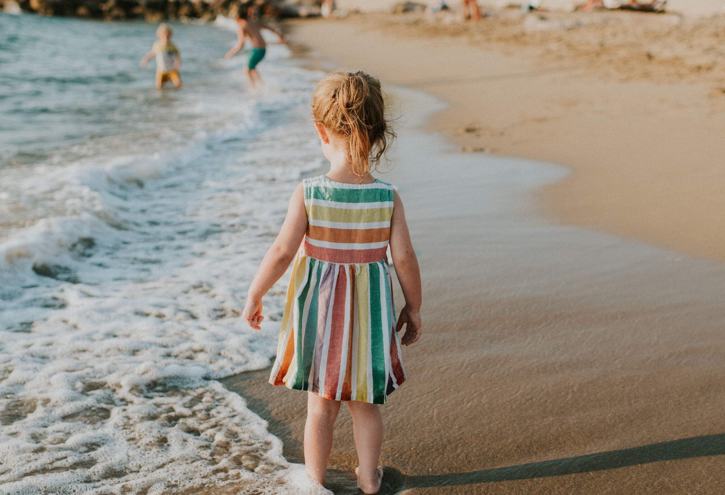A small girl wearing a colourful striped dress walking along the shore.