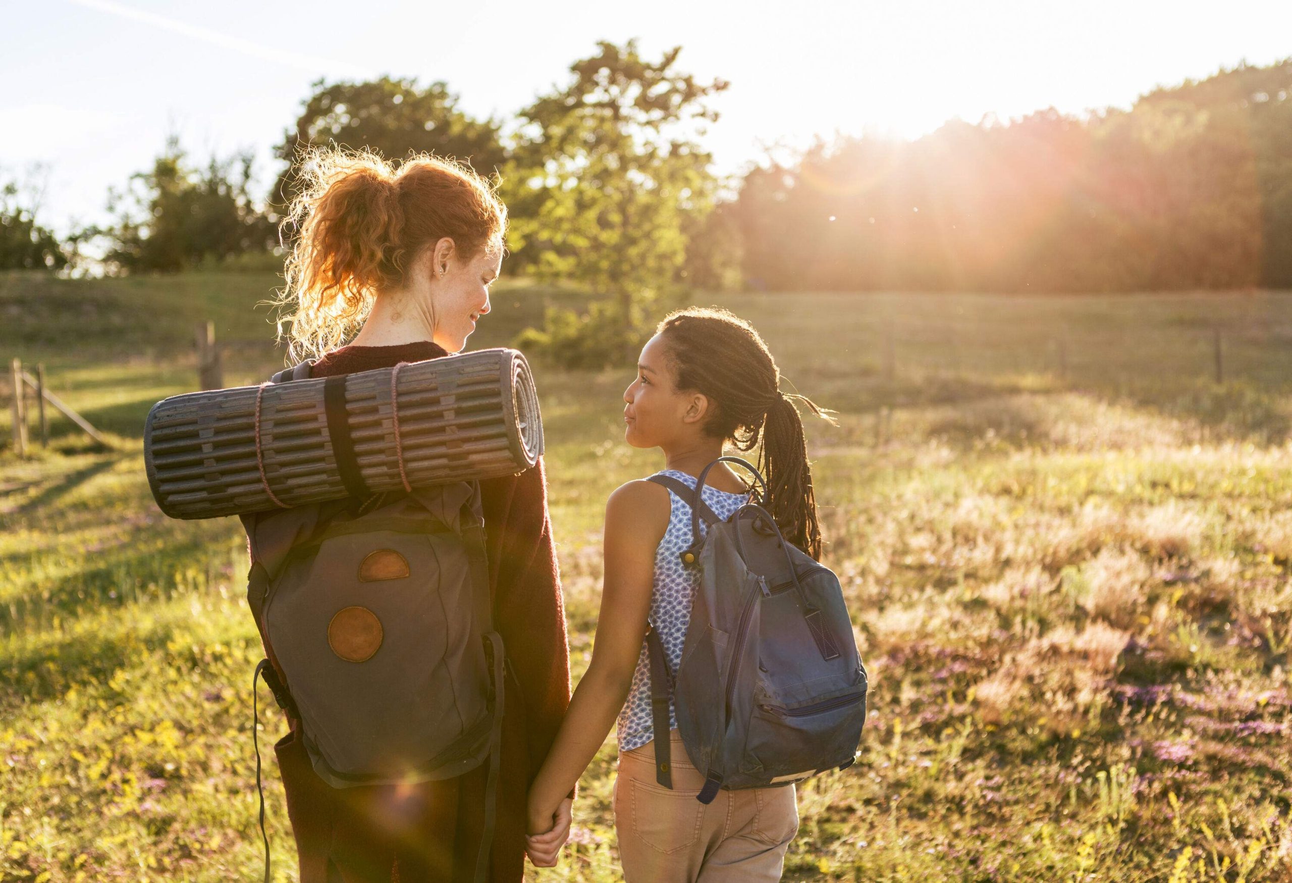 A woman and a young girl with backpacks walking across a grassland with the sun shining over the trees.