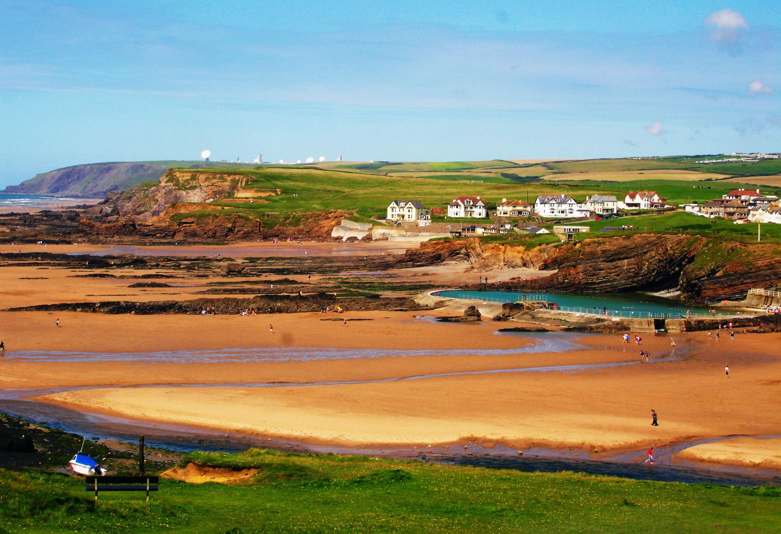A sandy stretch beach shore features a rock pool beneath the lush cliff with classic white houses.