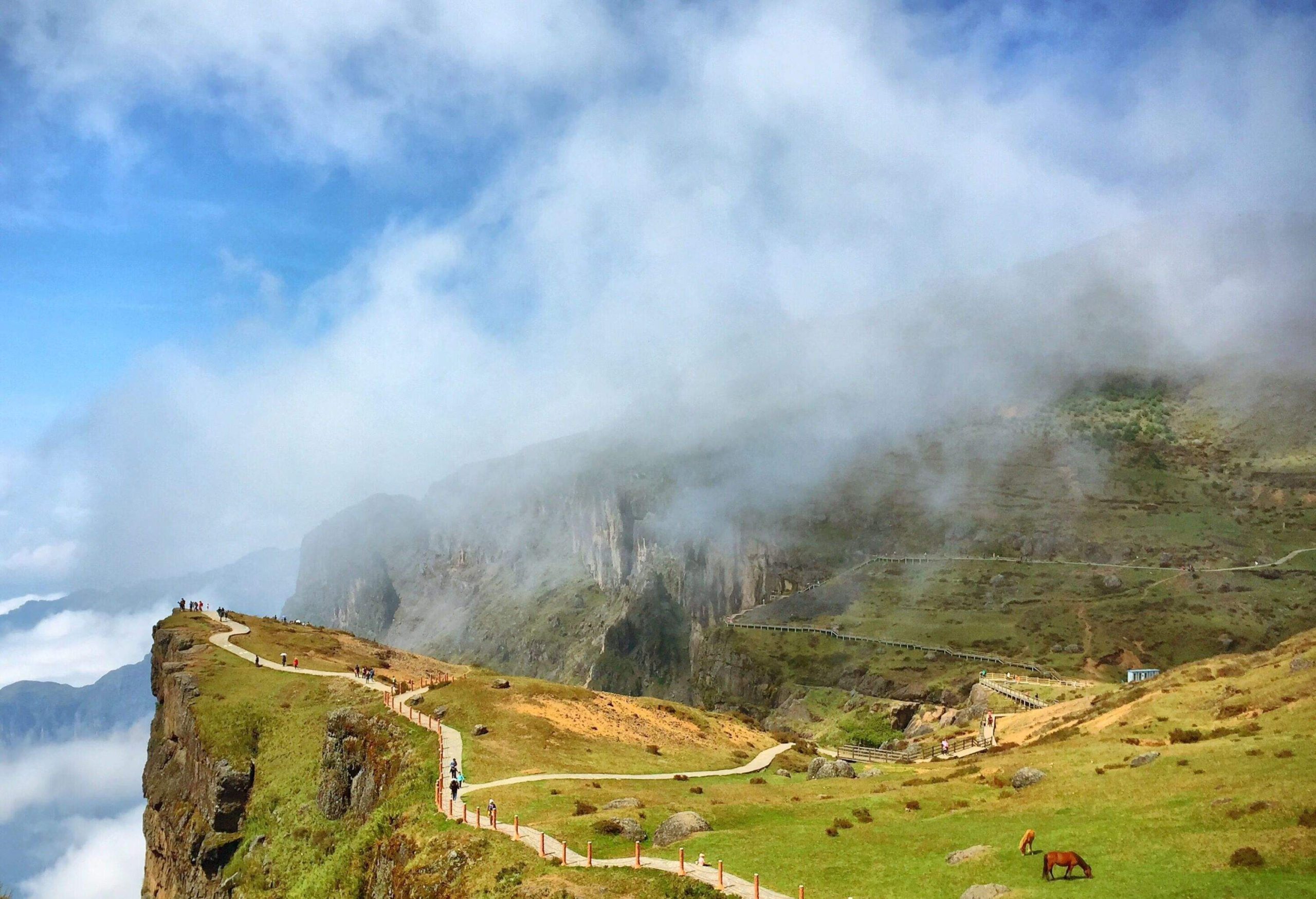 People walking on a paved trail towards a steep cliff with foggy hills in the distance.