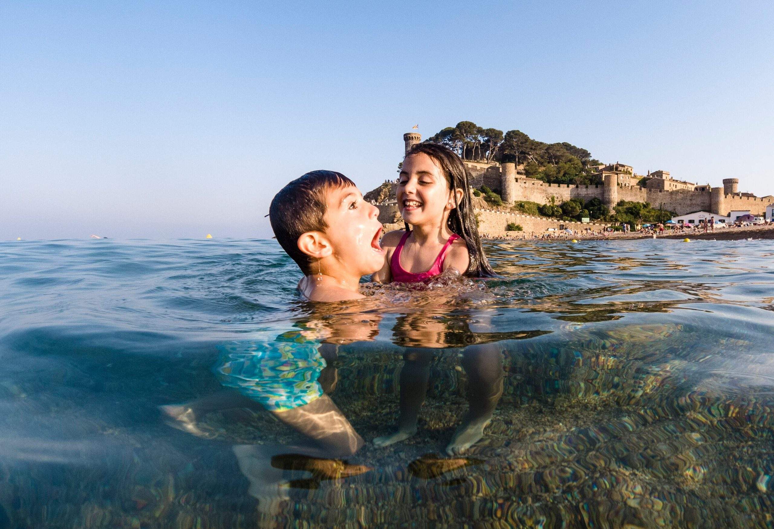 A couple of children playing in the sea with a fortress in the background.