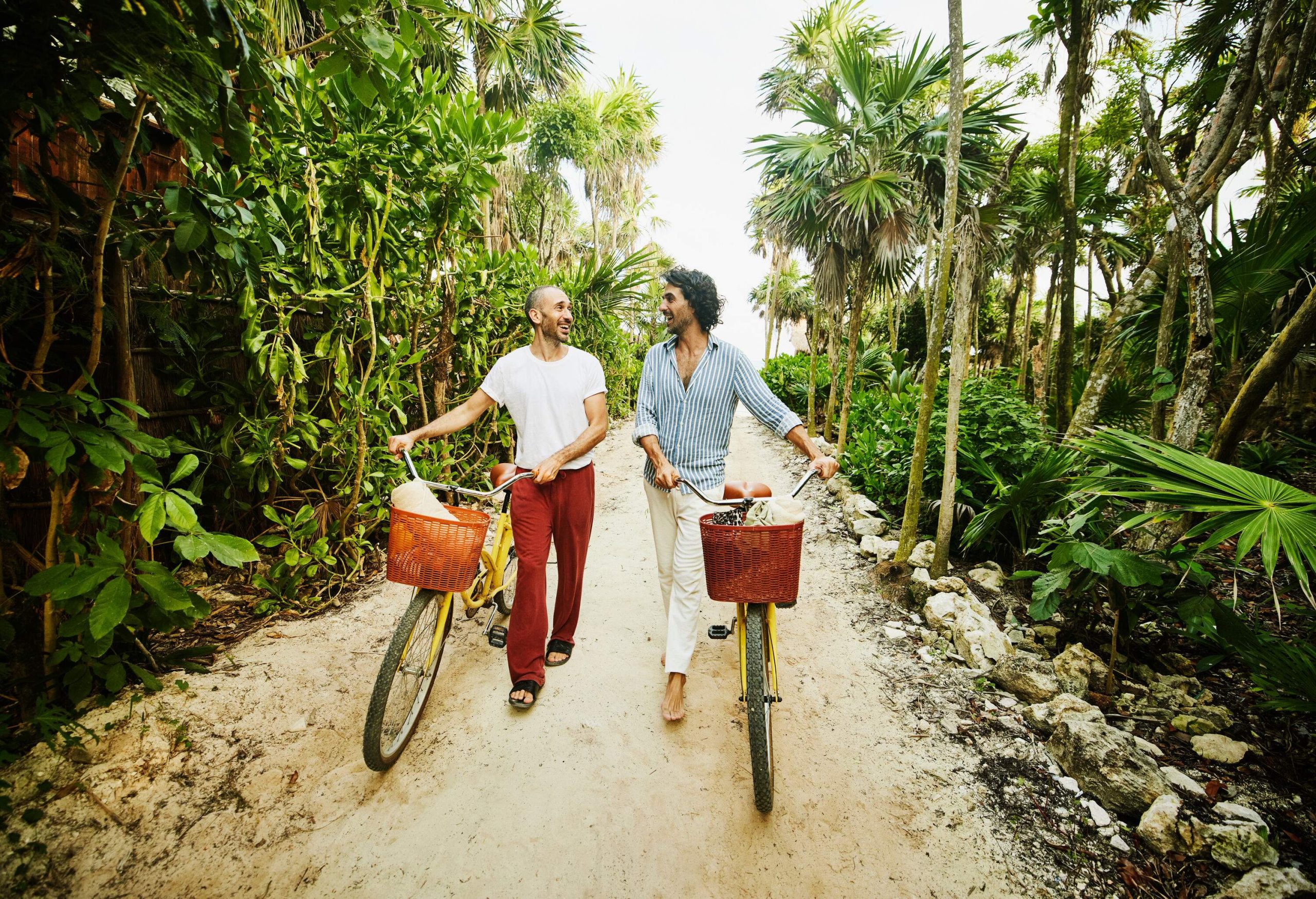 A laughing gay couple walking their bikes on a path surrounded by lush vegetation.