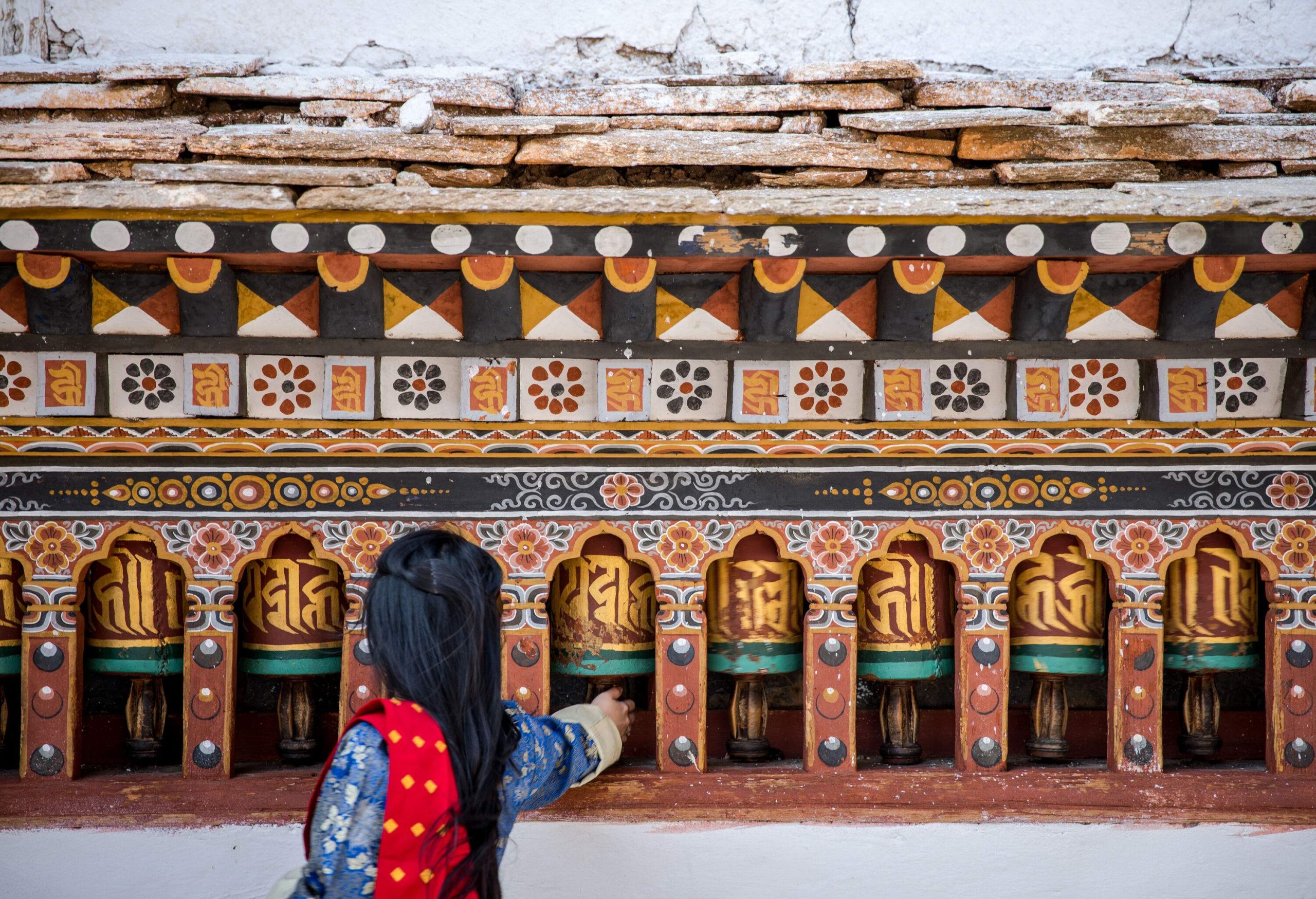 Gracefully adorned in a traditional Bhutanese dress called Kira, a woman delicately reaches out to touch one of the exquisitely painted, multi-coloured Bhutan prayer bells.