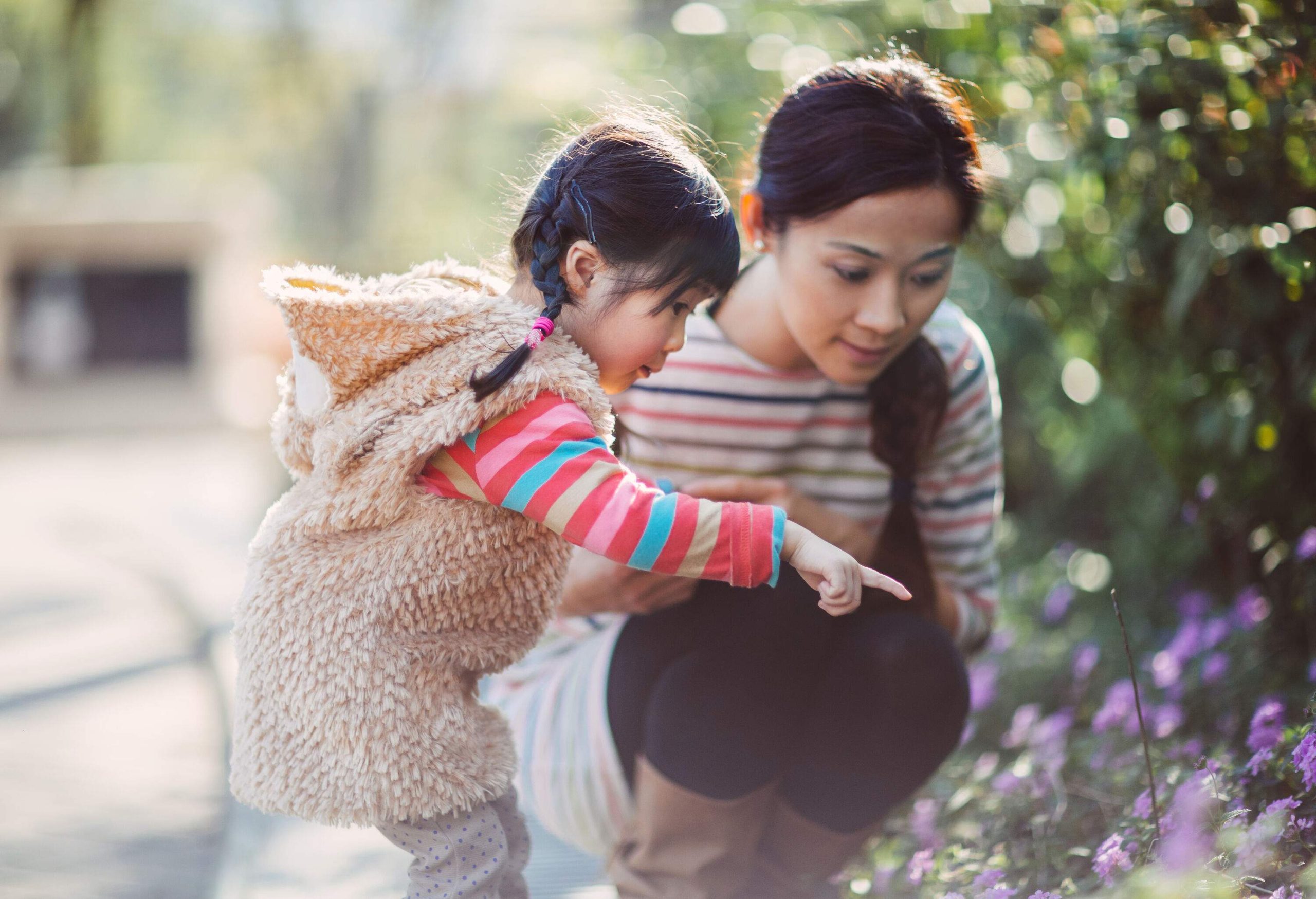 A girl in a colourful furry hoodie points at the sidewalk flowers as her mother looks on.