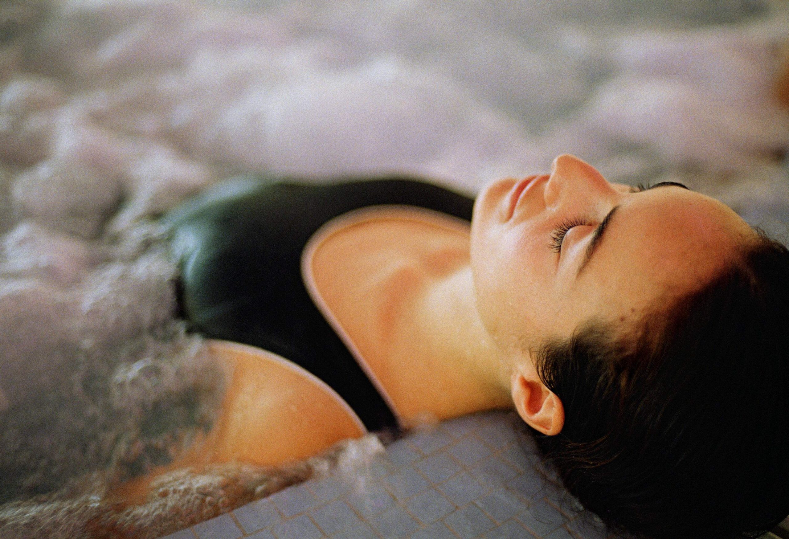 A woman in a black swimsuit relaxes in a hot tub with her eyes closed.