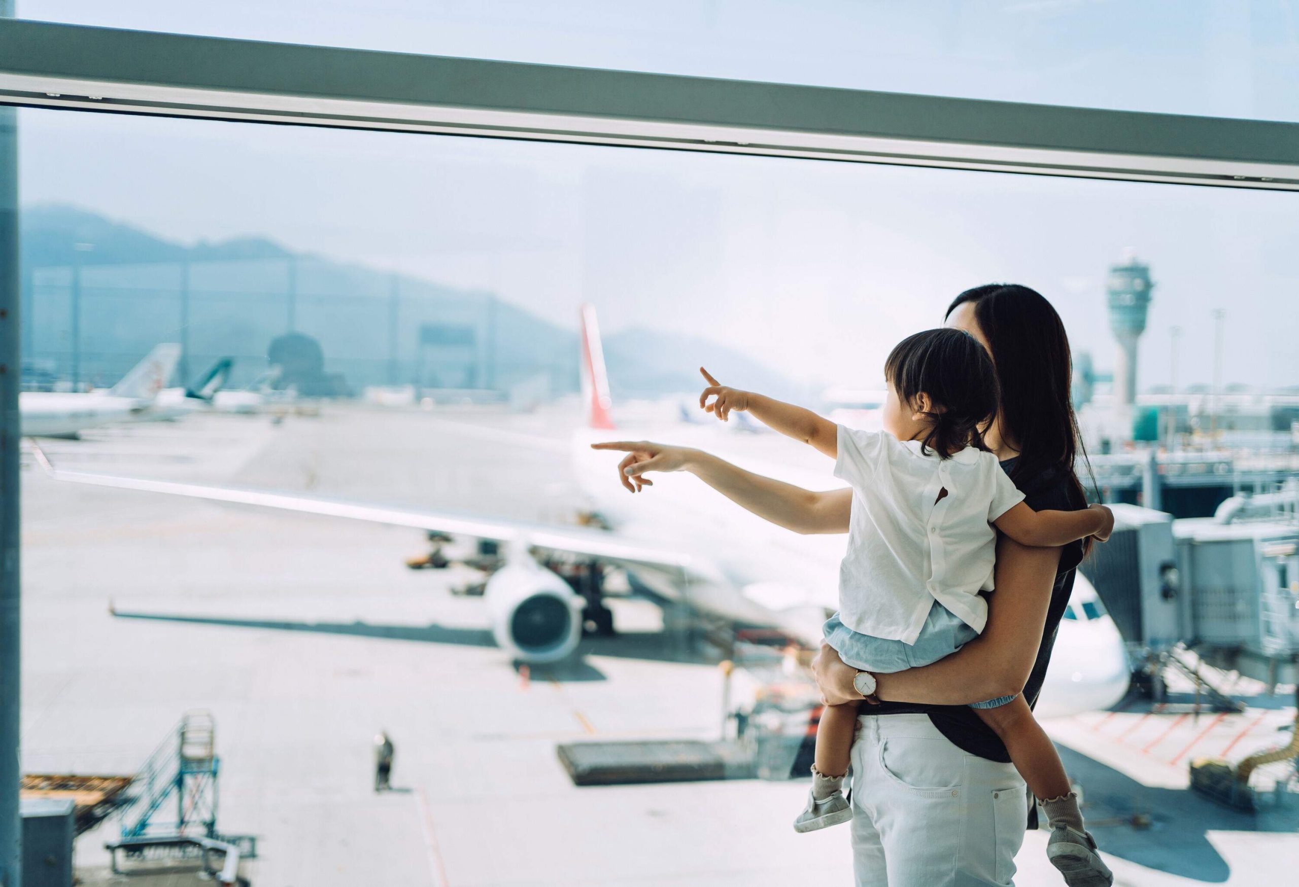 A mother carries her toddler while standing next to a window and pointing at the planes.