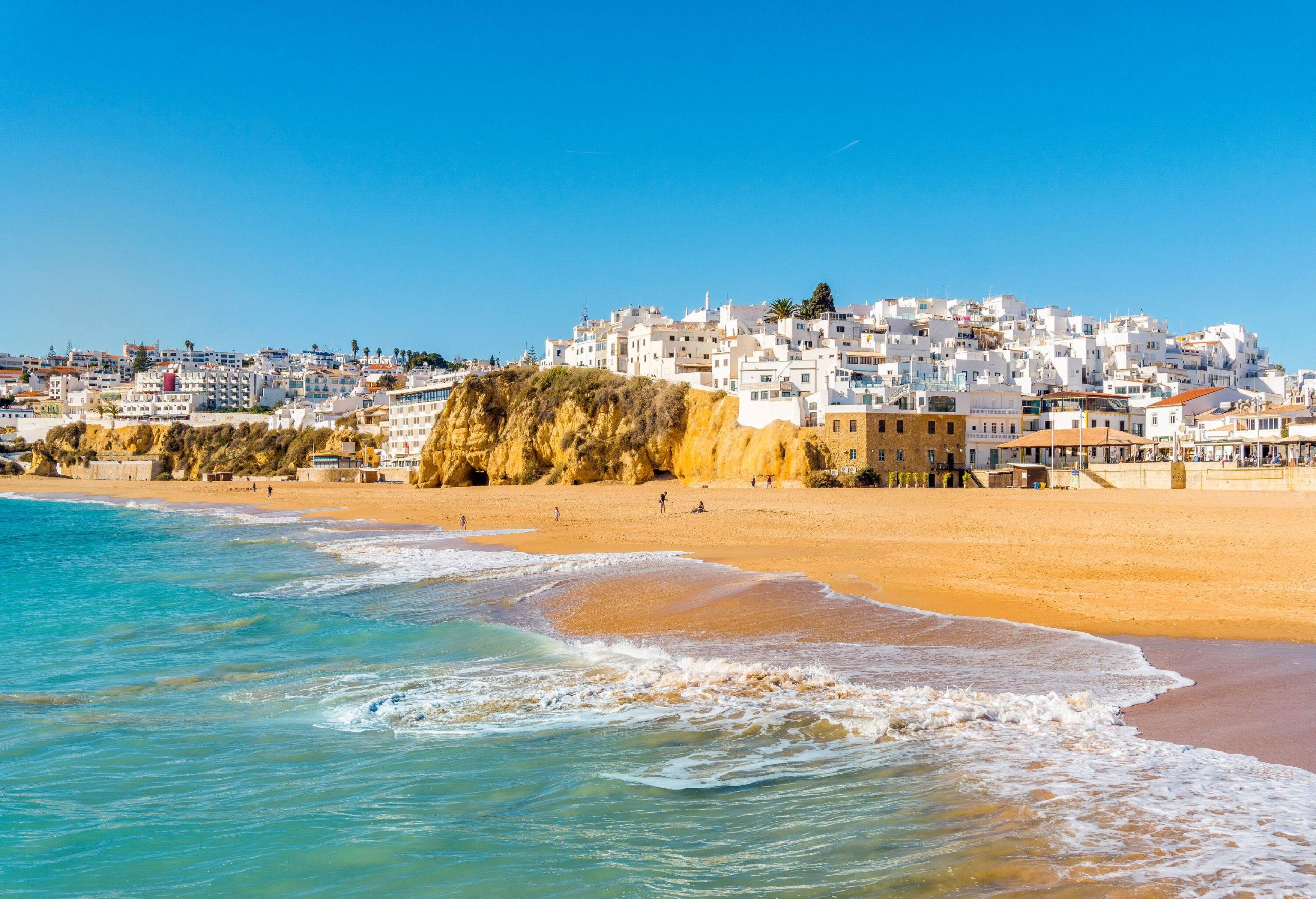 A sandy beach lined with white houses and buildings against a clear blue sky.
