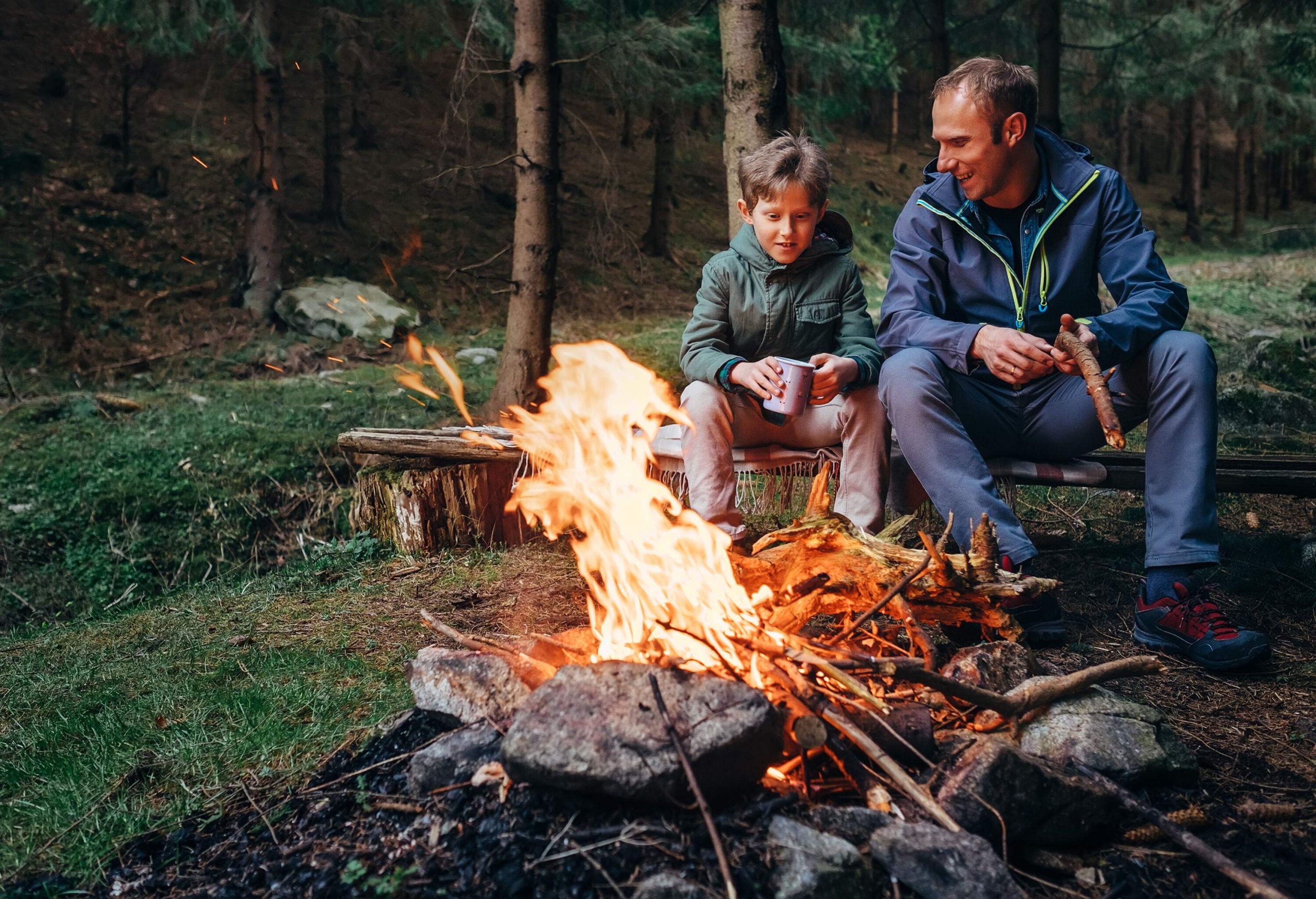 Father and son converse while sitting in front of a bonfire in the woods.