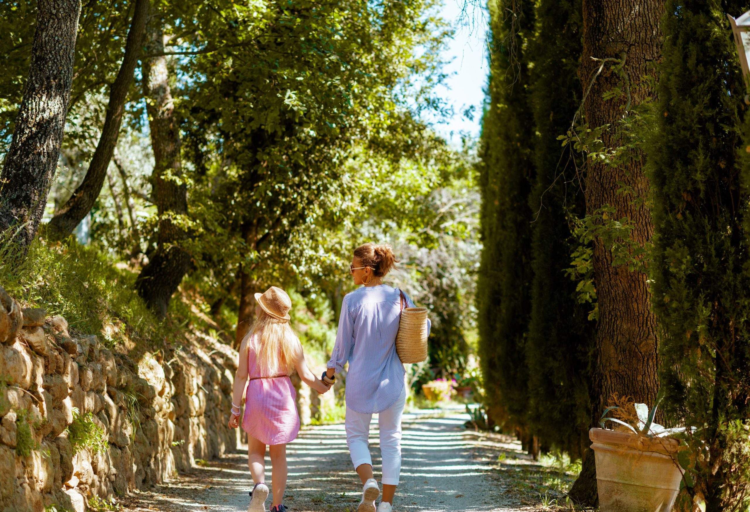 A woman and a girl hold hands as they stroll along a path in the shade of trees.