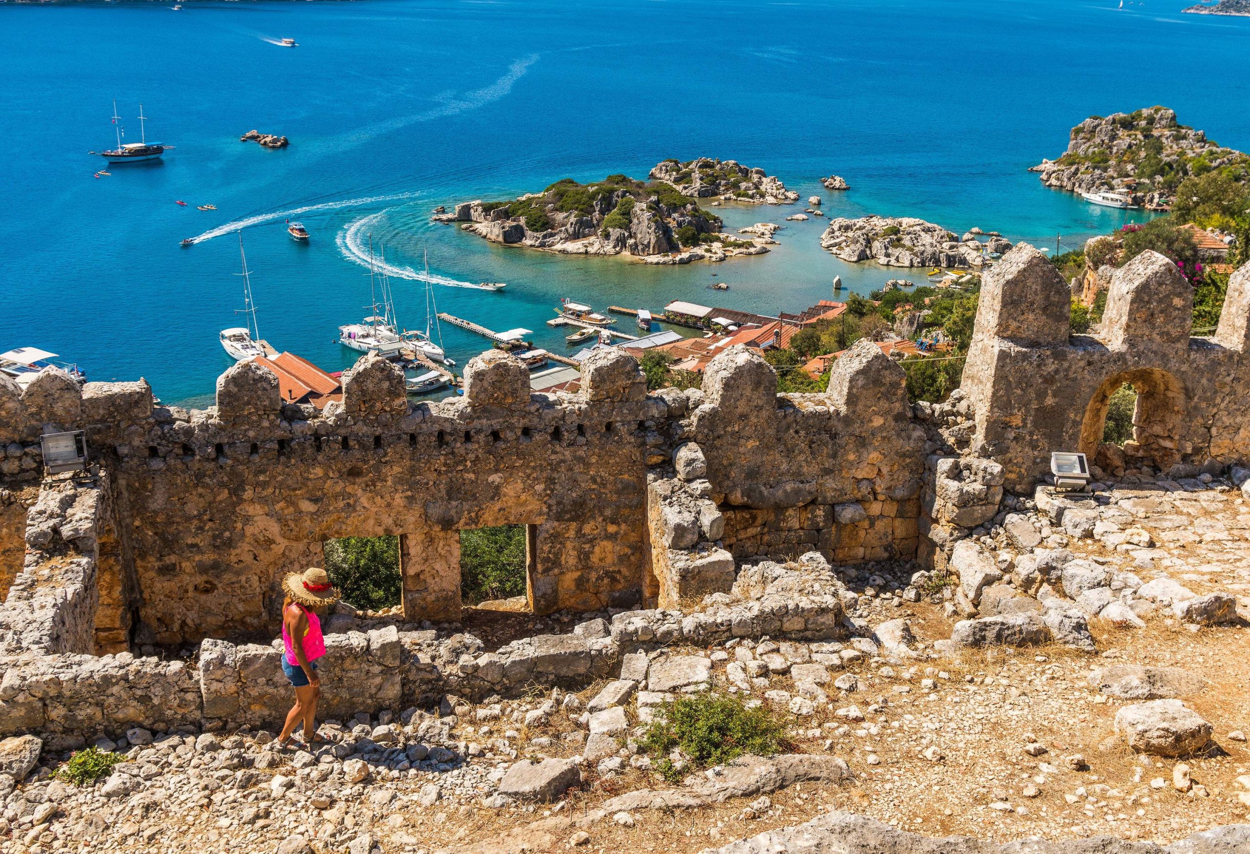 A female tourist wanders around a ruined castle overlooking several boats and the blue bay.
