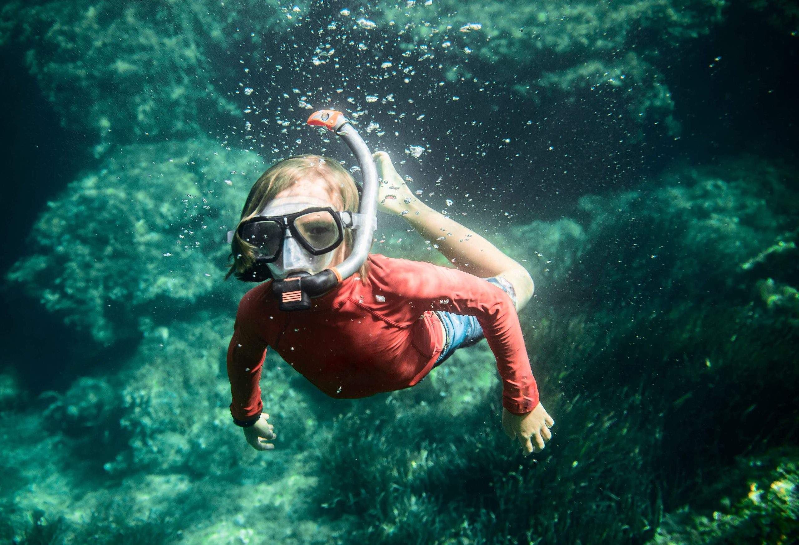 A kid in a red rash guard dives into the ocean wearing a snorkelling gear.