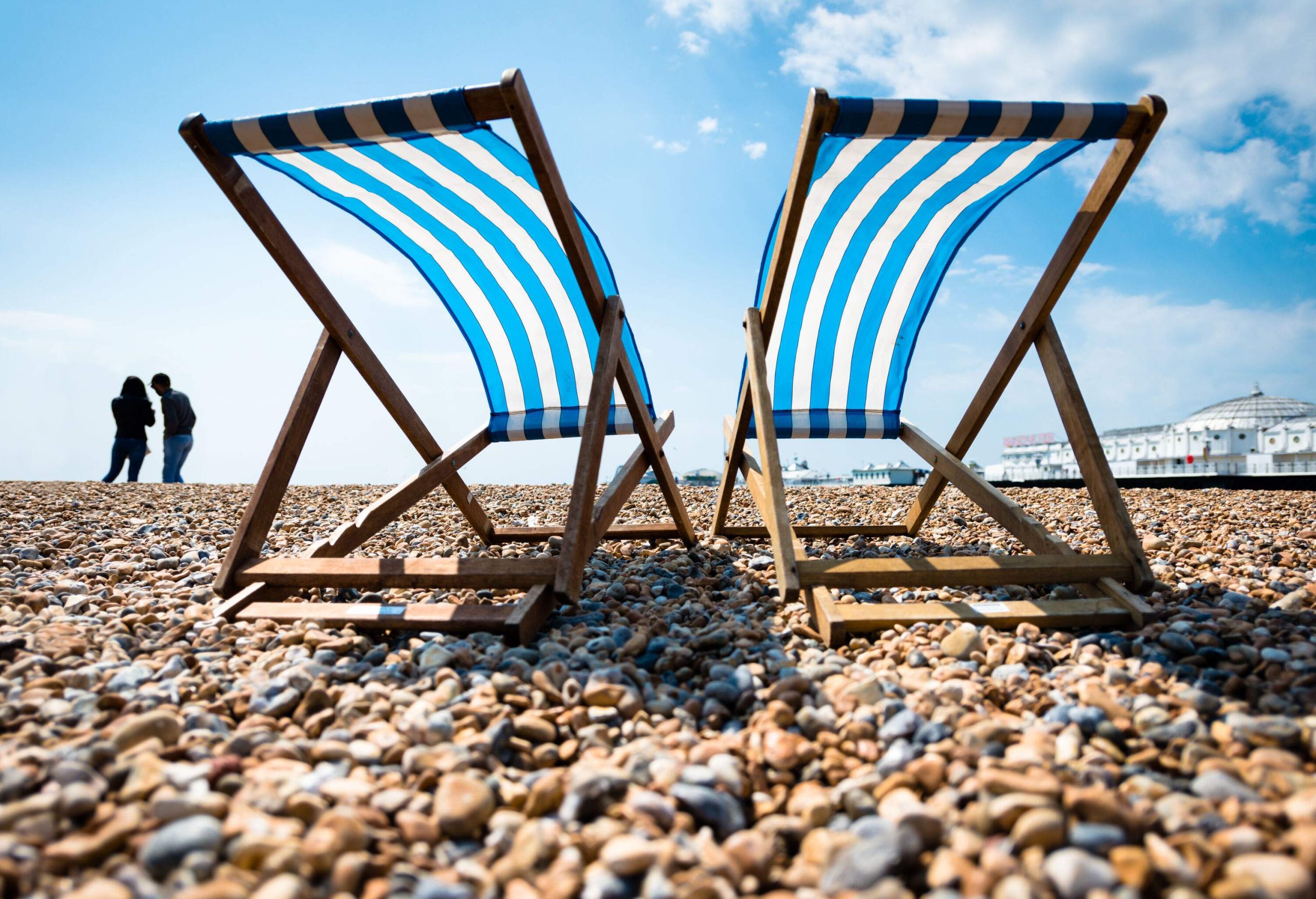 Two deck chairs set on a pebbly beach and a silhouette of a couple standing in the distance.