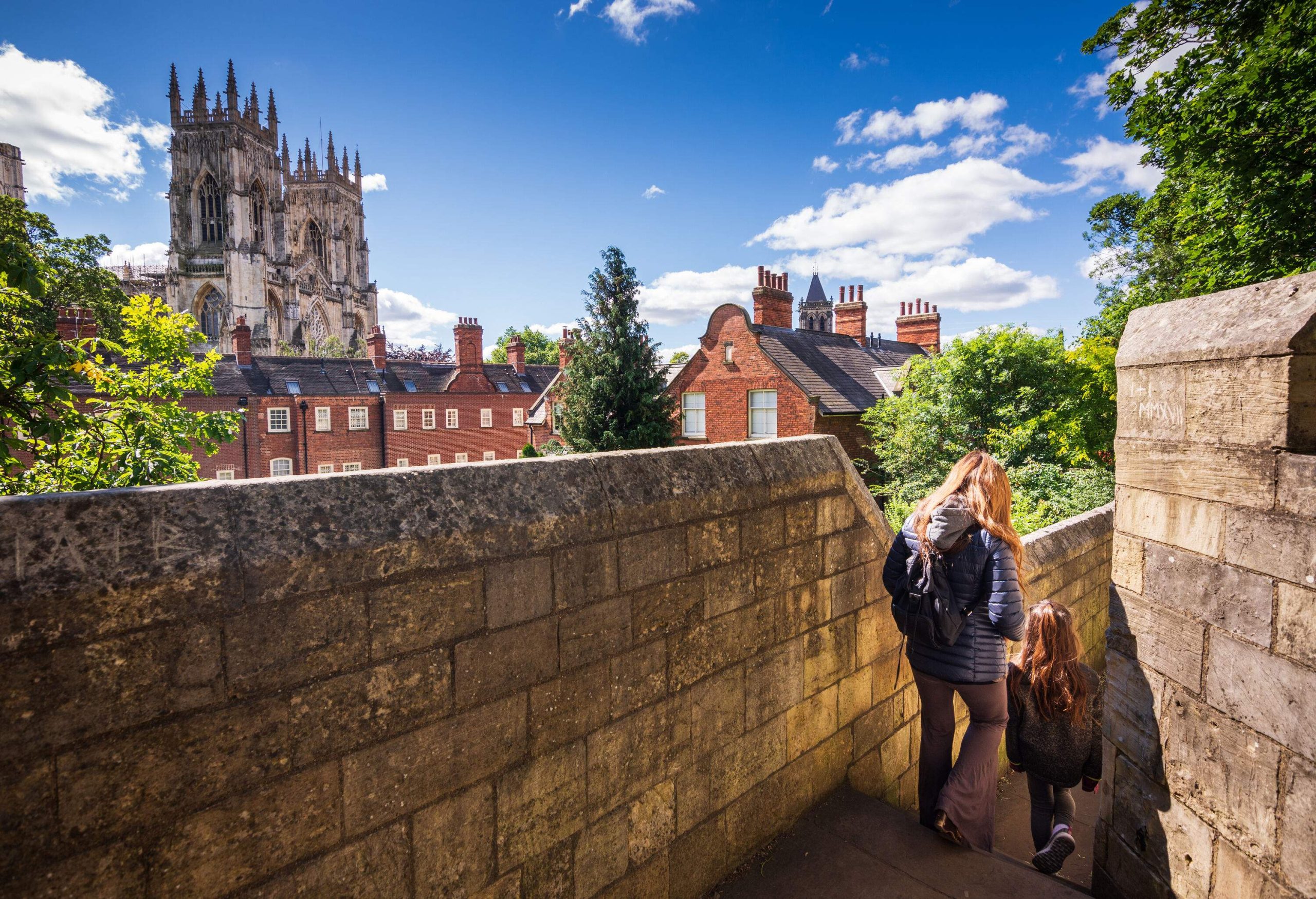 A mother and a daughter go down the stairs of a city wall with a view of church towers.