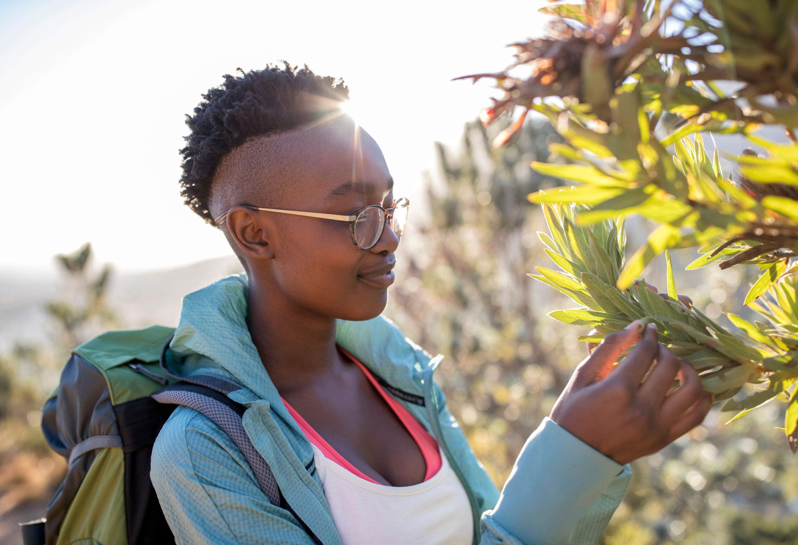 A short-haired hiker with glasses smiles while touching some tree leaves.