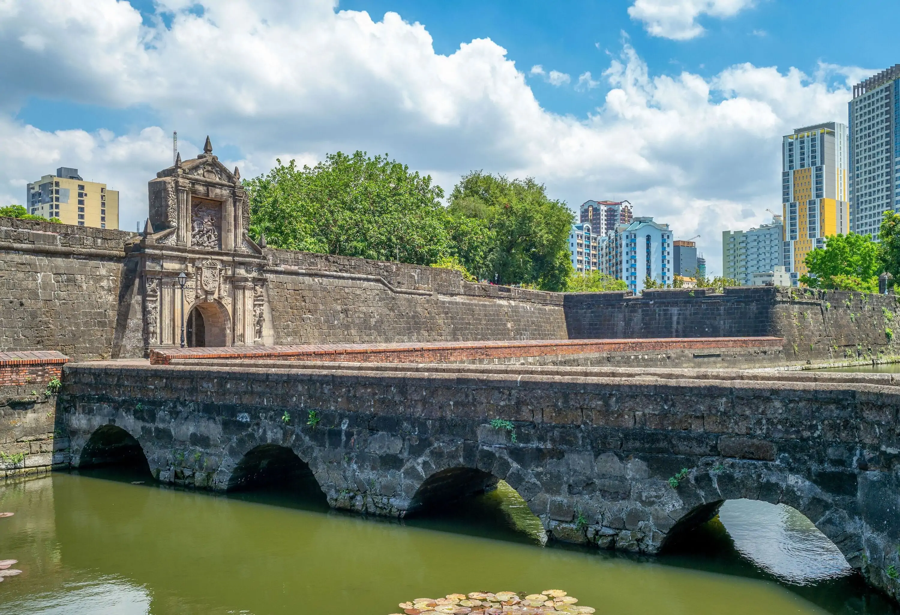 An arch bridge leading to the main gate of a fortress, with modern structures rising in the background.