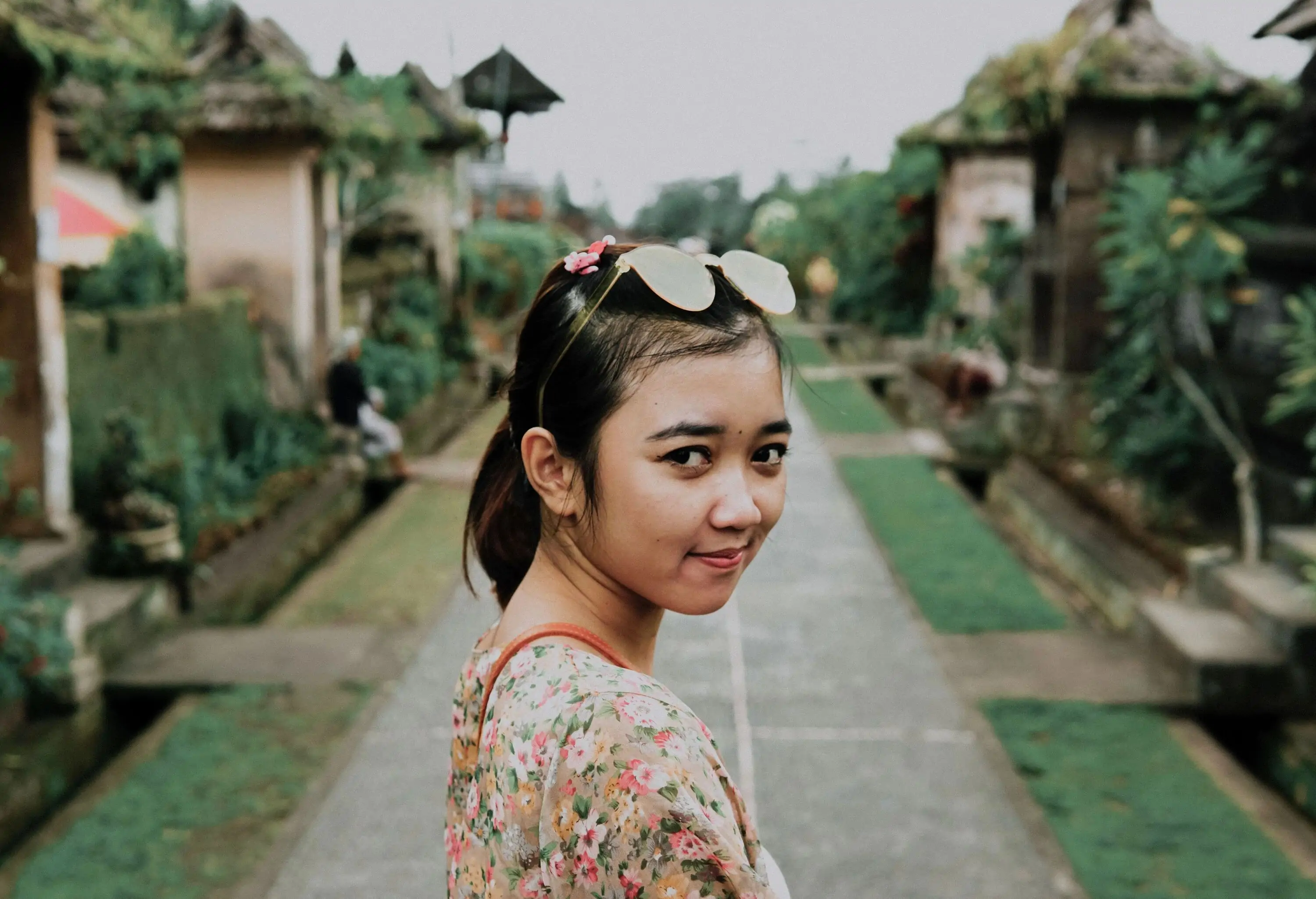A woman in a floral t-shirt with sunglass rests on her head against a blurred lush background.