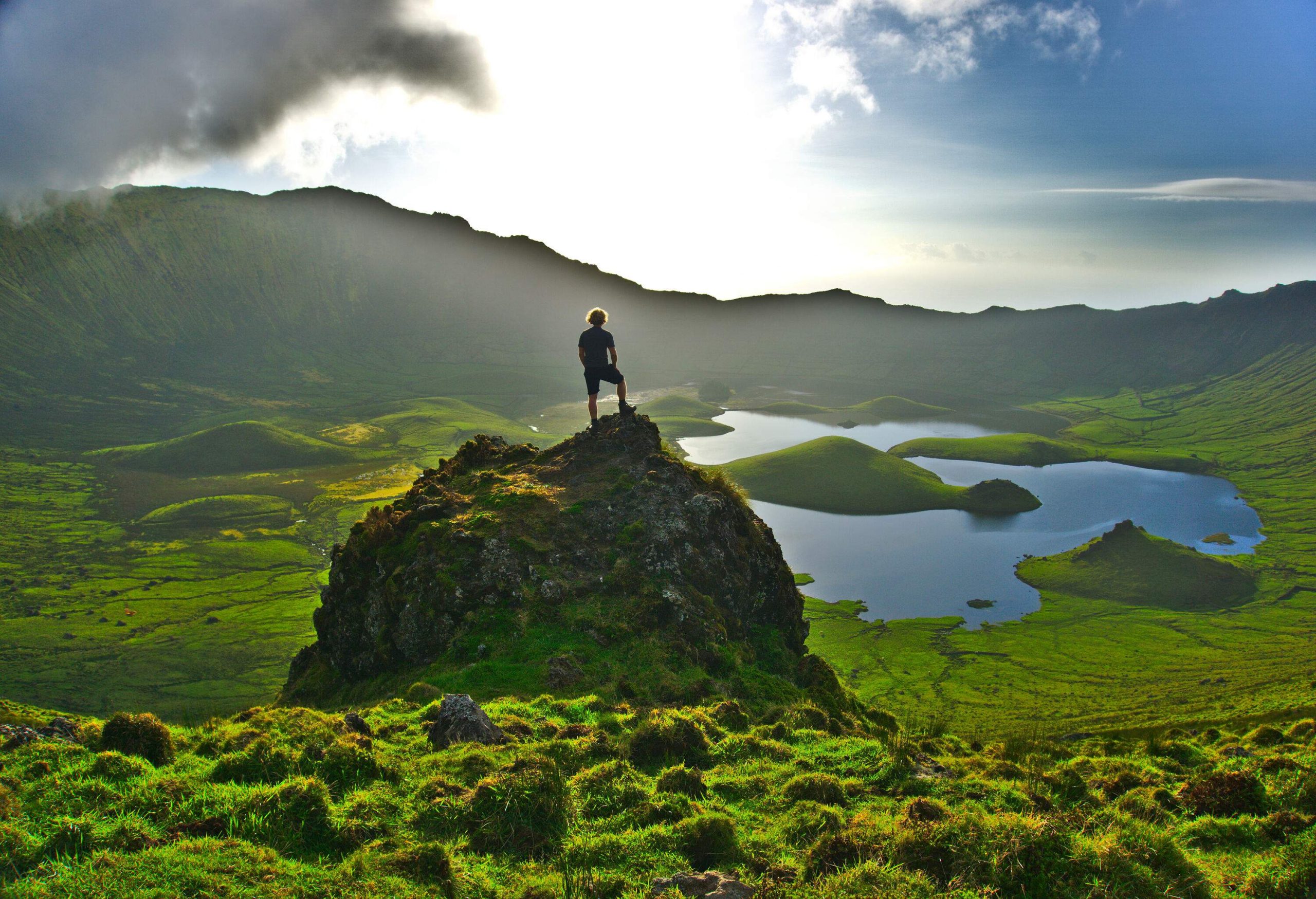 A person standing on a grass-covered boulder while staring at the crater and mountains in the distance.