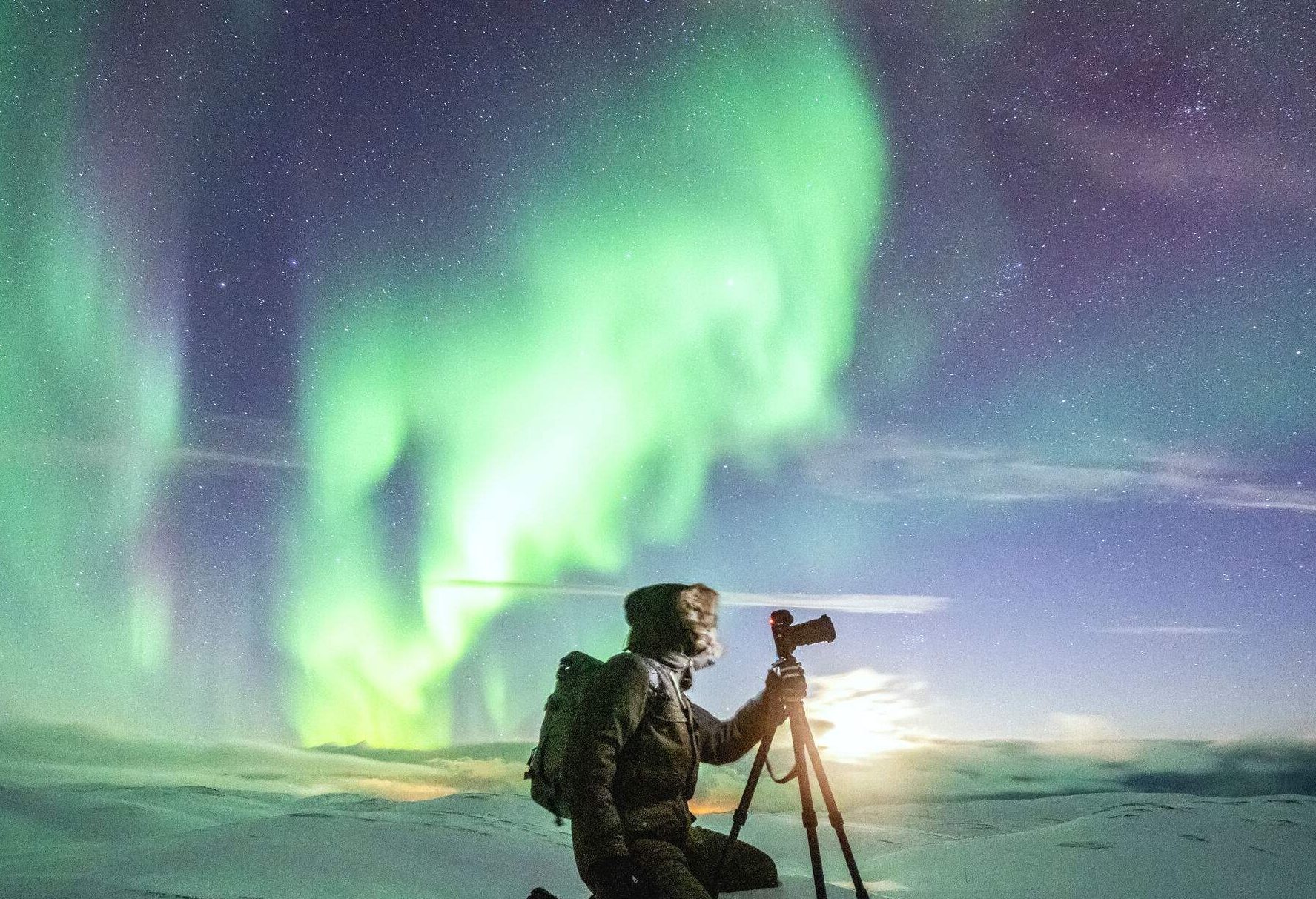 A photographer takes pictures of the Northern green lights dazzling in the sky with bended knees on his tripod.