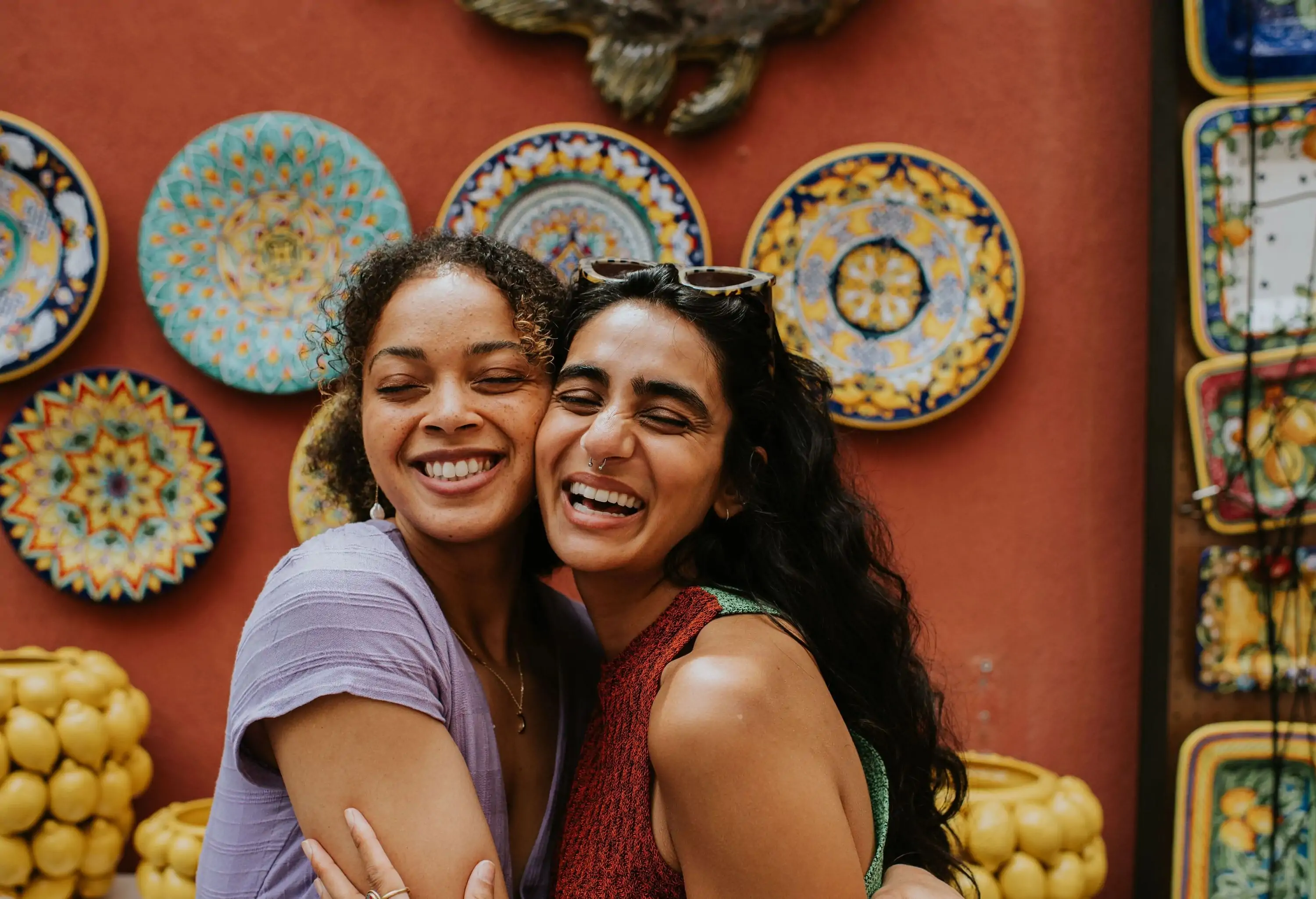 Two young female friends shop in Positano, Italy, famous for its colourful ceramics. They pose together outside a red wall, with wall mounted plates and vases. The woman affectionately embrace as they smile confidently at the camera.