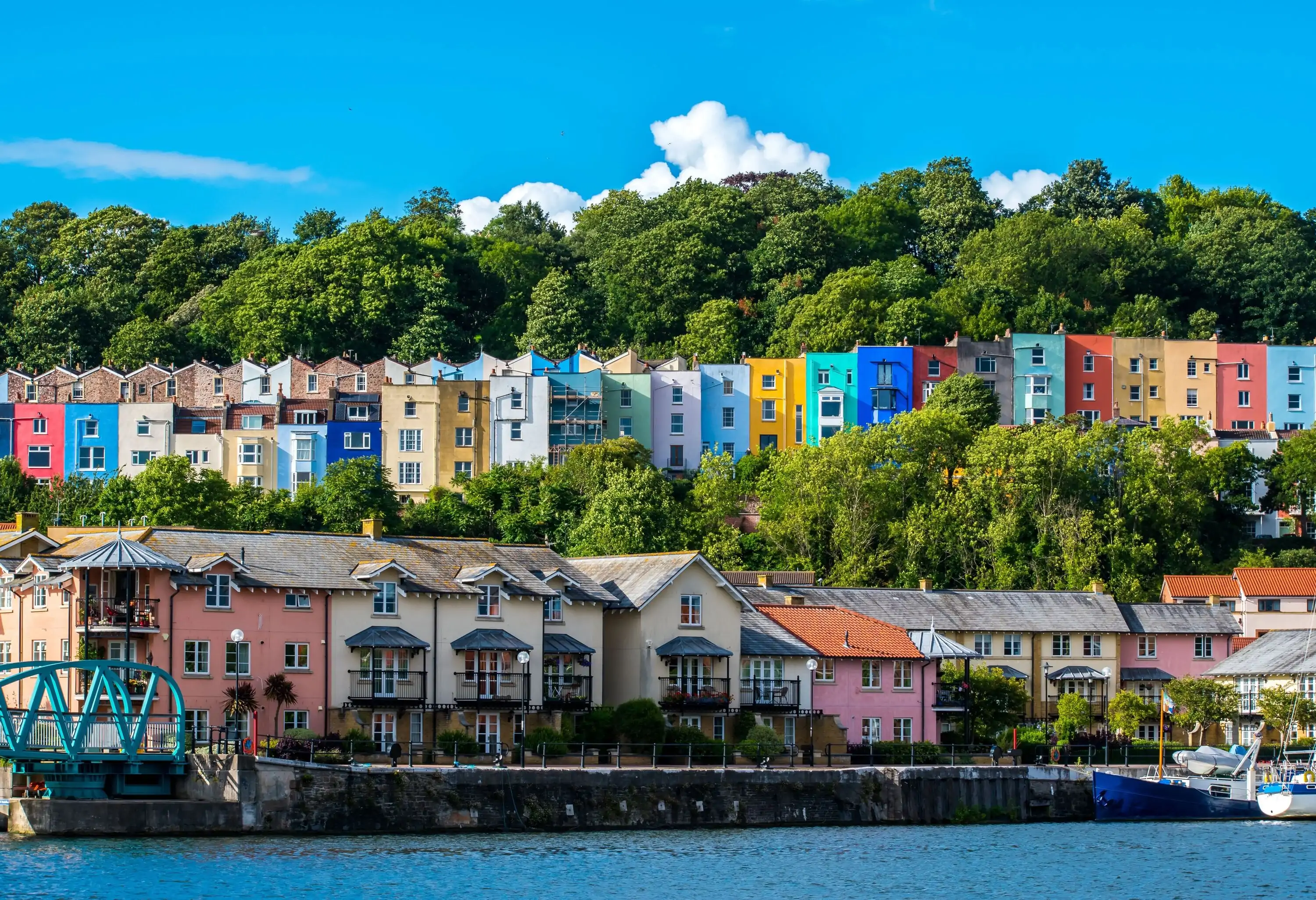 Line of colourful townhouses, overlooking a body of water, with big trees in the back and a row of homes in front.
