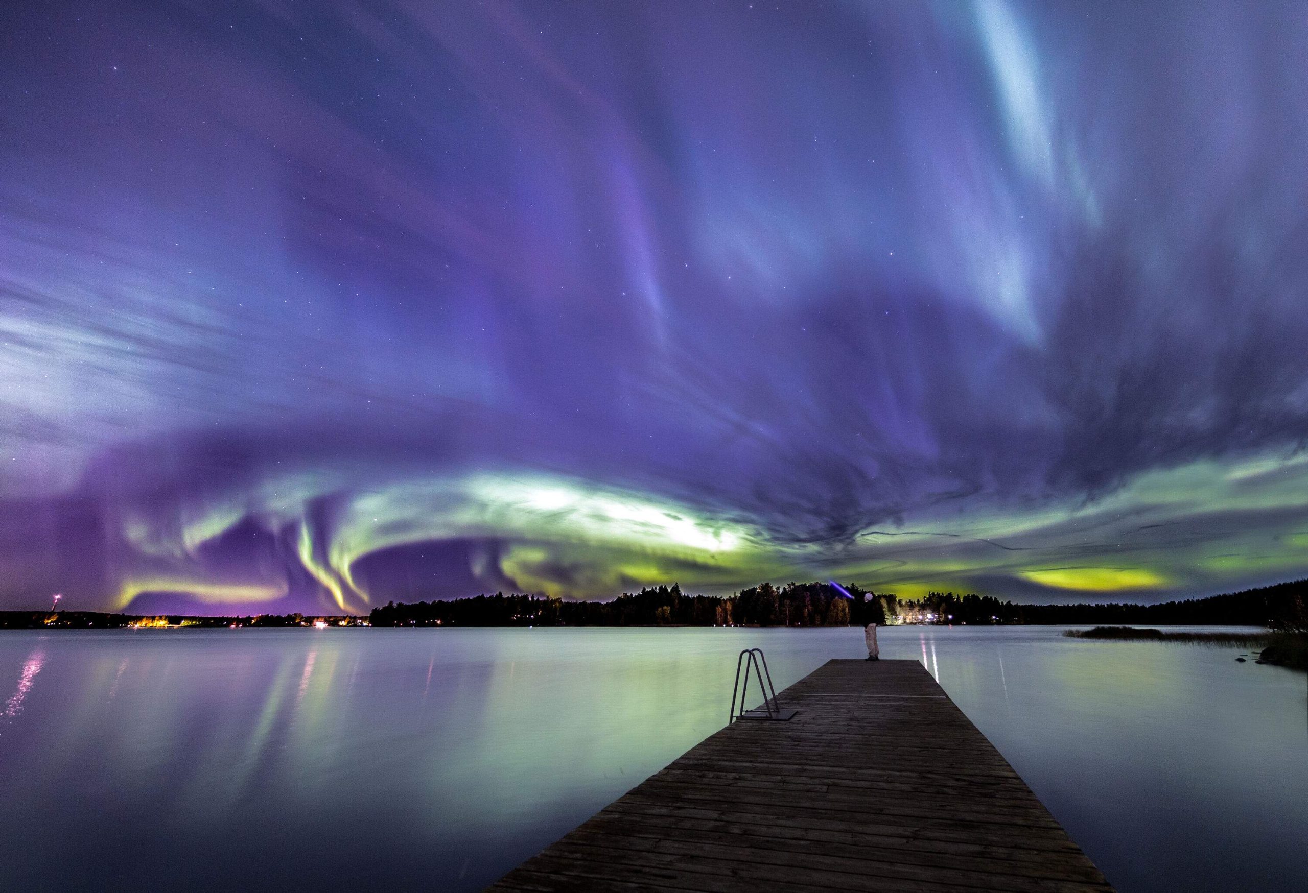 A man standing at the end of a boardwalk, watching the northern lights swirl across the sky.