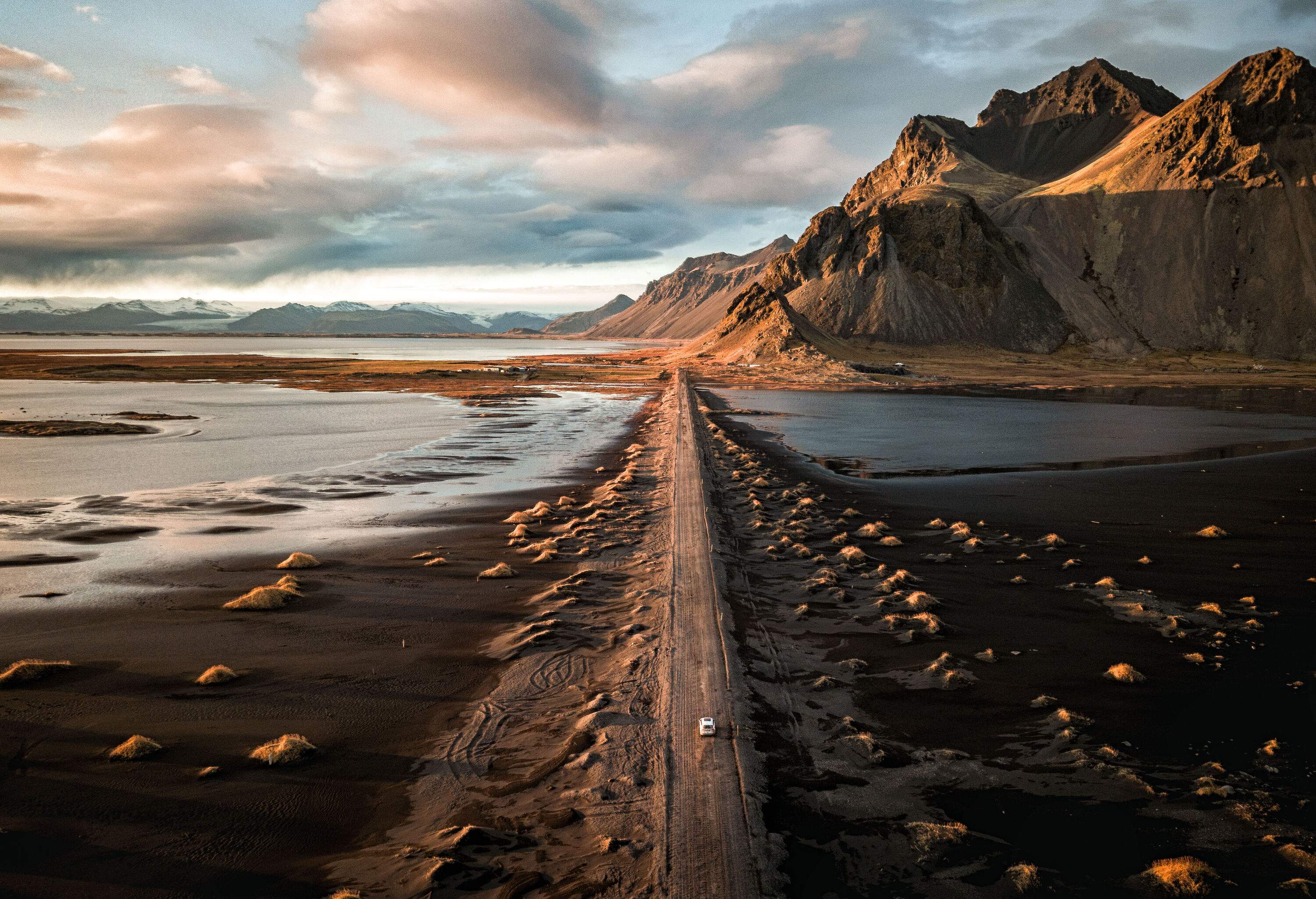 A white car travelling a narrow road in the middle of a rugged black sand valley surrounded by steep pointed mountains.