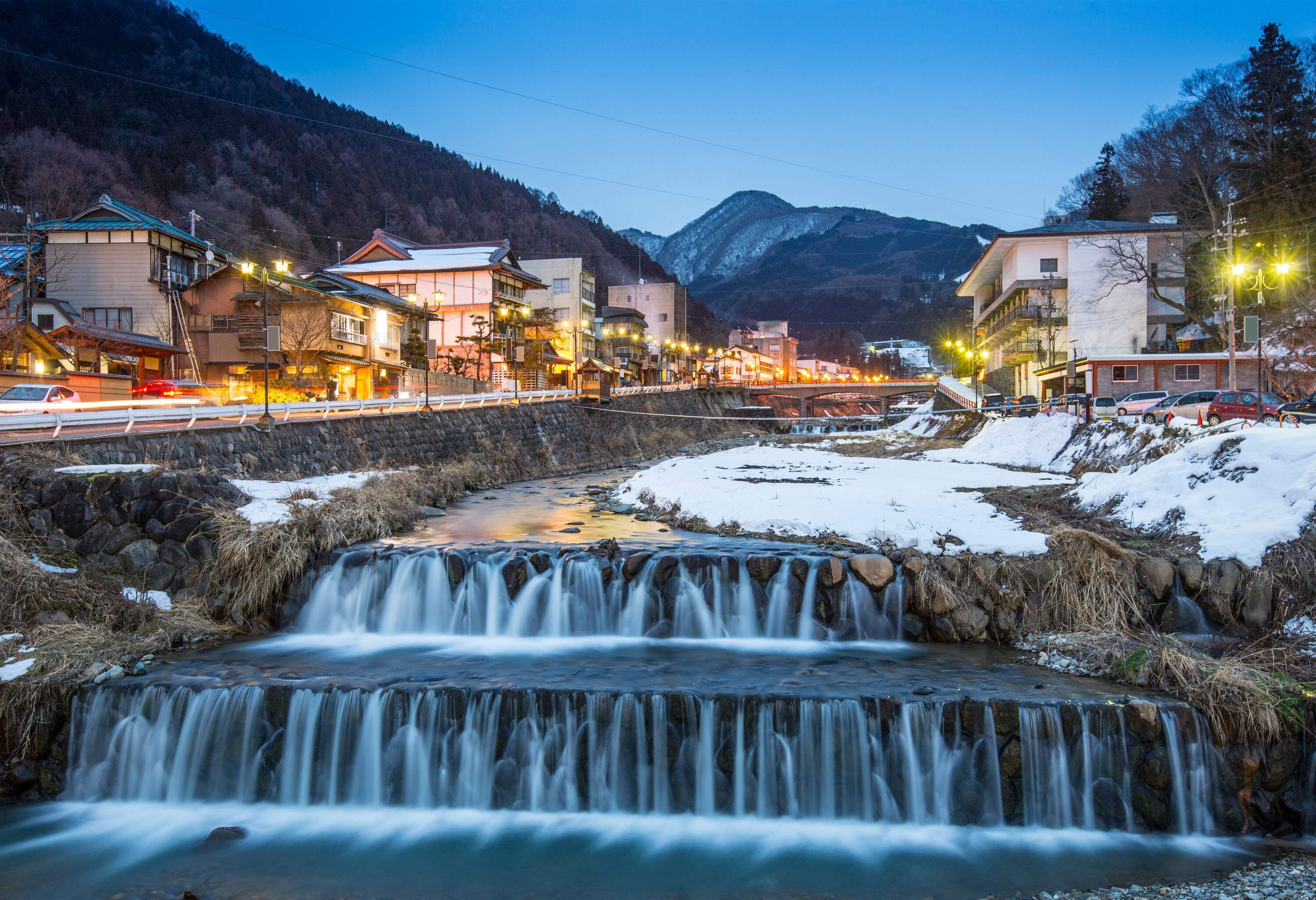 A terraced river lined with the bustling road and buildings beneath the forested hill captured at twilight.