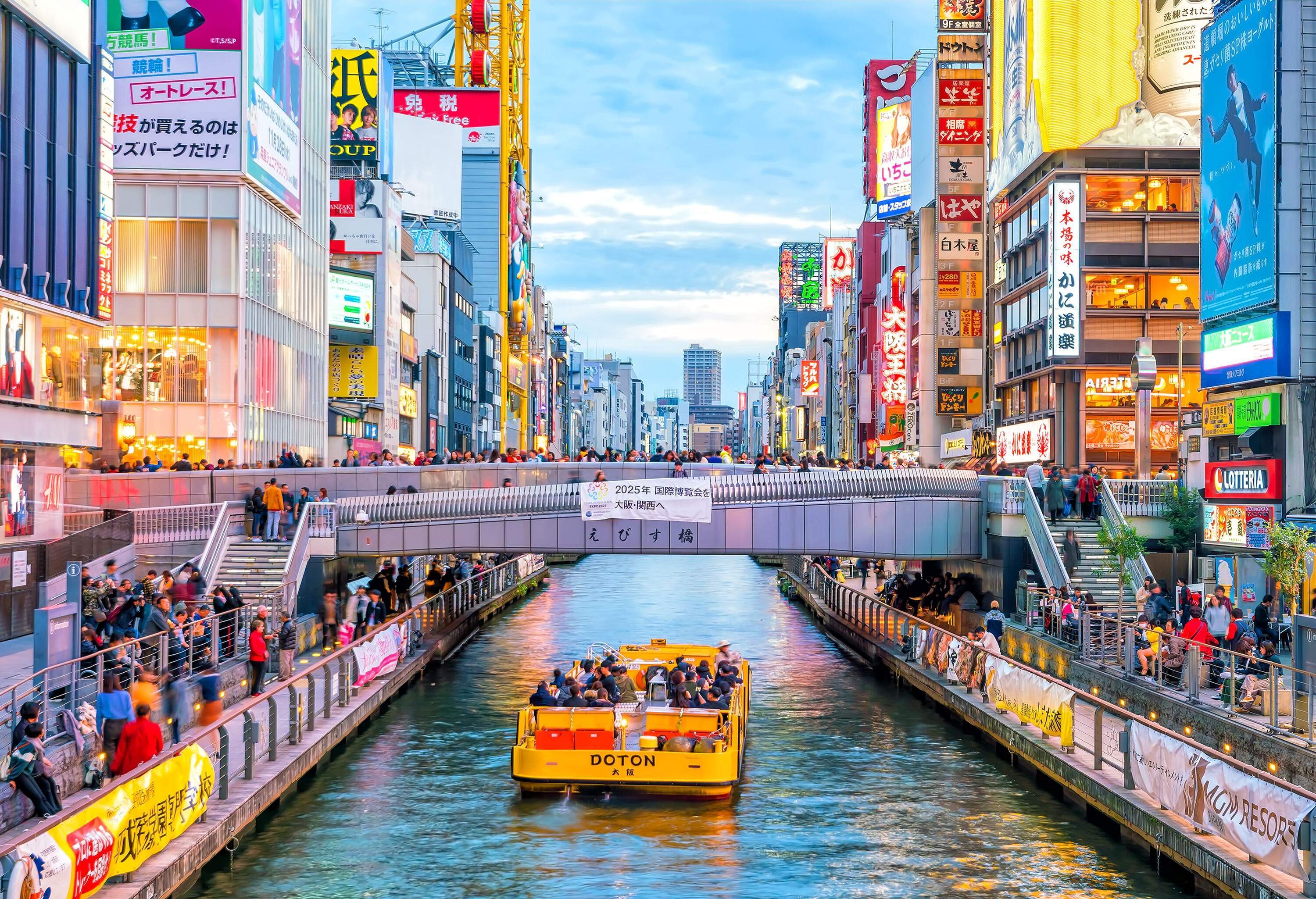 A yellow ferry gracefully glides beneath a bridge, while the bustling banks teem with pedestrians strolling amidst vibrant advertisements.