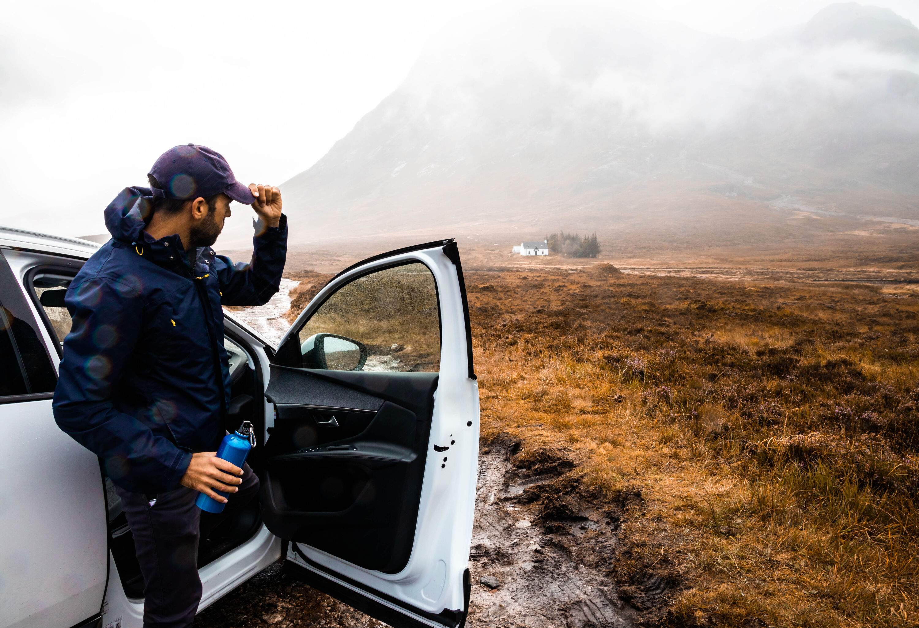 Man clad in a blue windbreaker emerges from a white car, holding a flask and looking at rugged landscape on a cloudy day