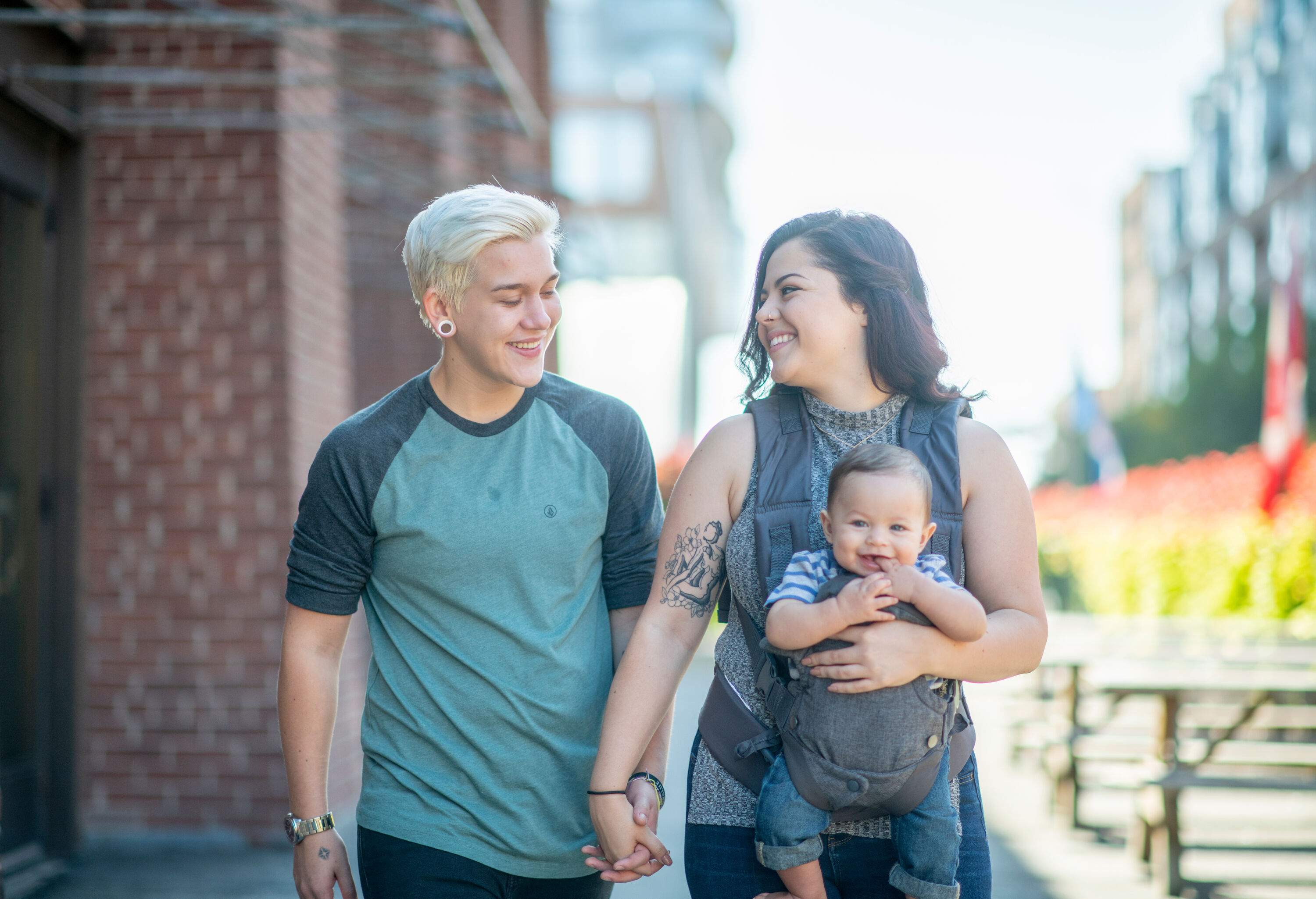 A lesbian pair walking down the street carrying a baby in a carrier.