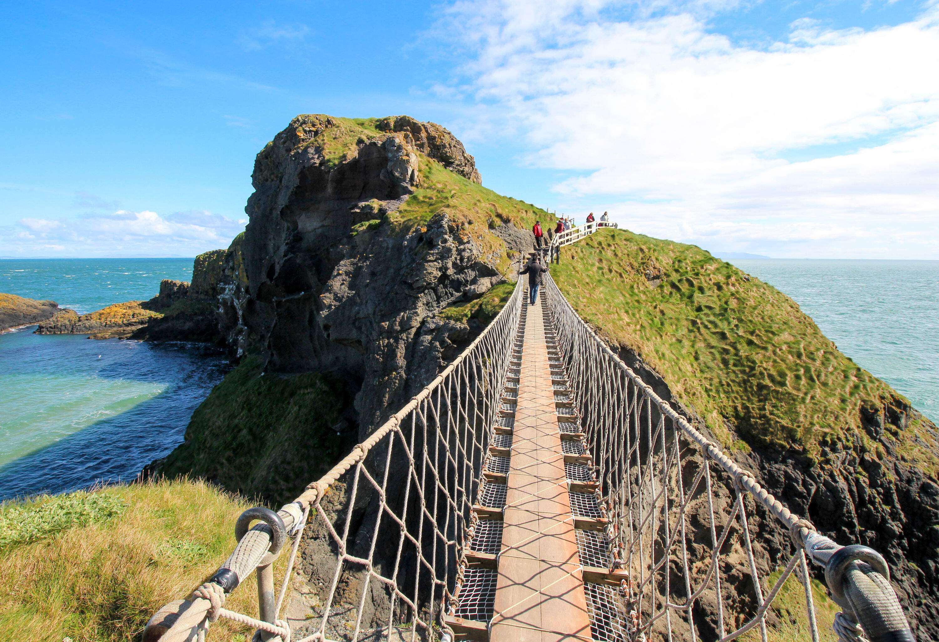 A long, narrow rope bridge leads to lush, jagged cliffs overlooking the sea.