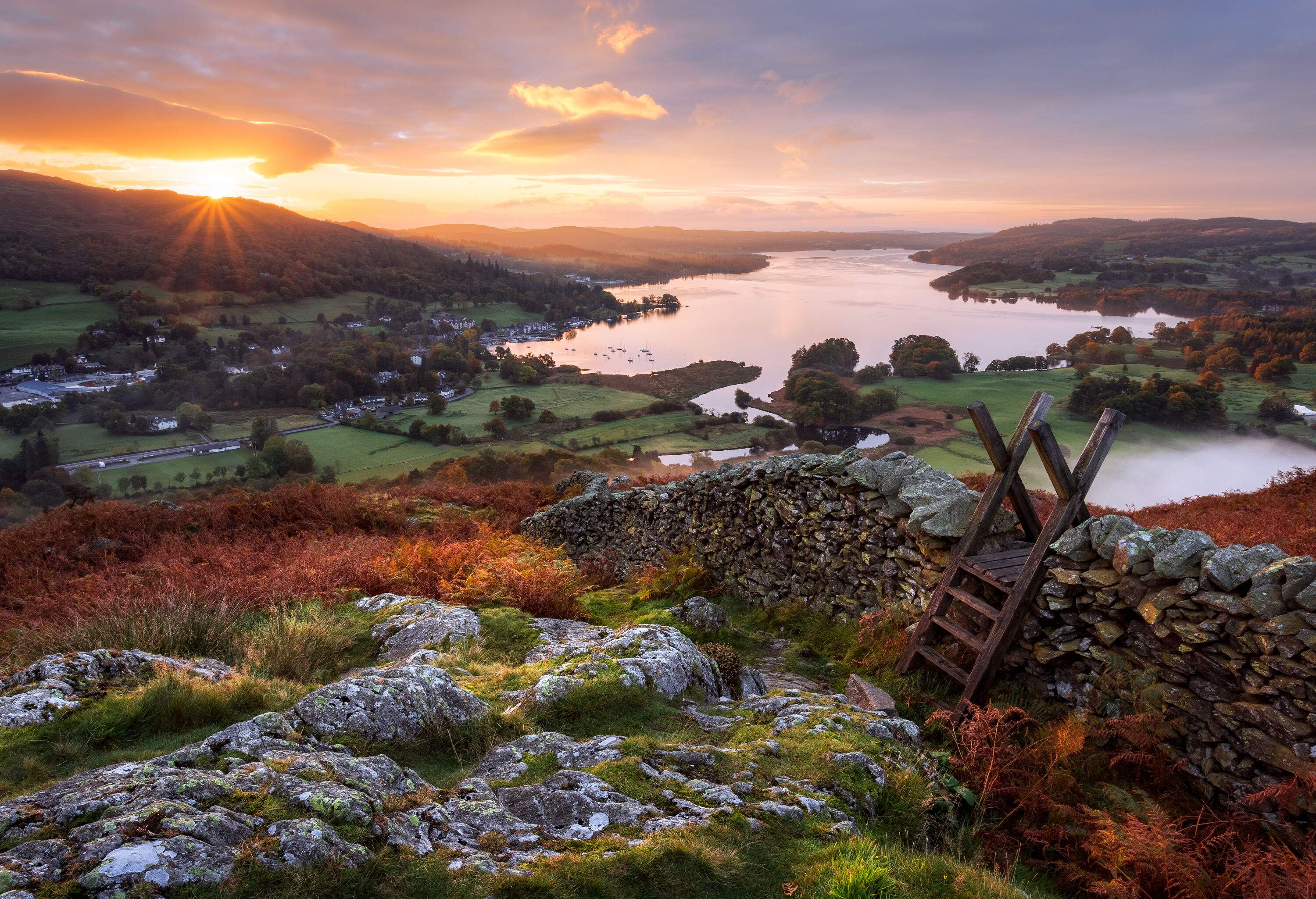 A stone wall with a ladder on a hill with overlooking views of a lakeside village and the setting sun.