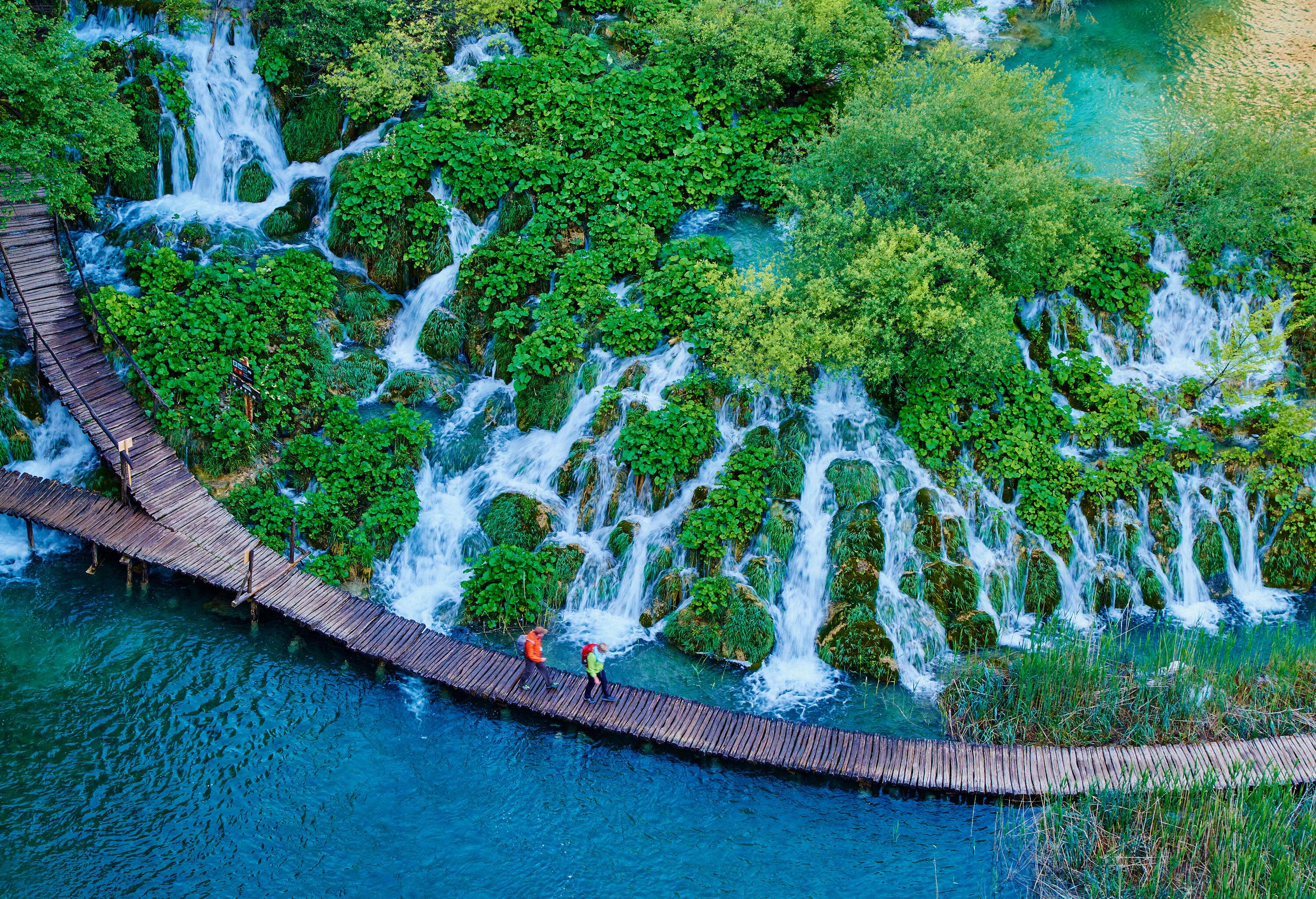 Two hikers walk on a wooden walkway across a stream beside a cascade of small interconnected waterfalls on rocks covered in green vegetation.