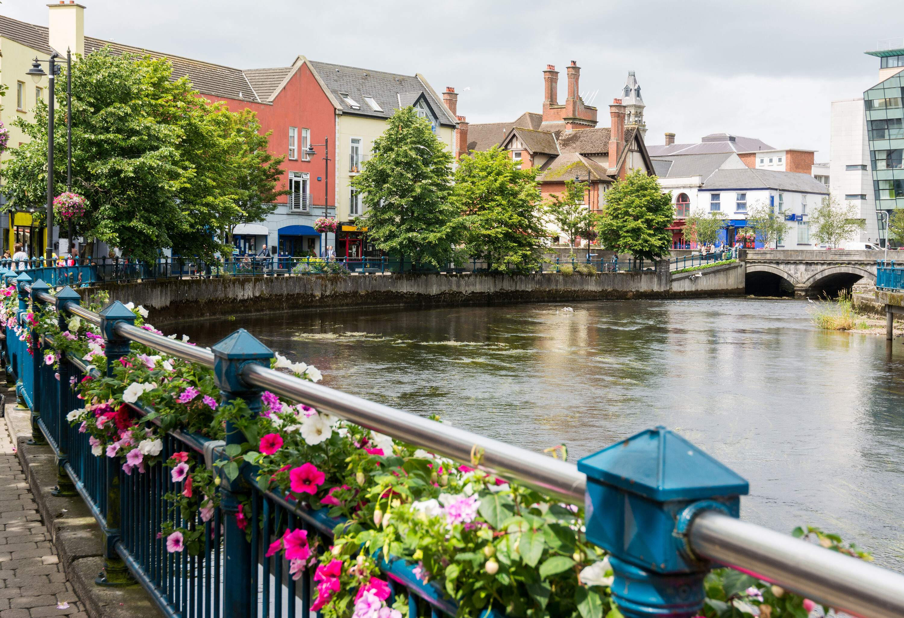 Blue-painted wrought iron fence ornamented with colourful flowers guards a promenade around a broad canal with trees.