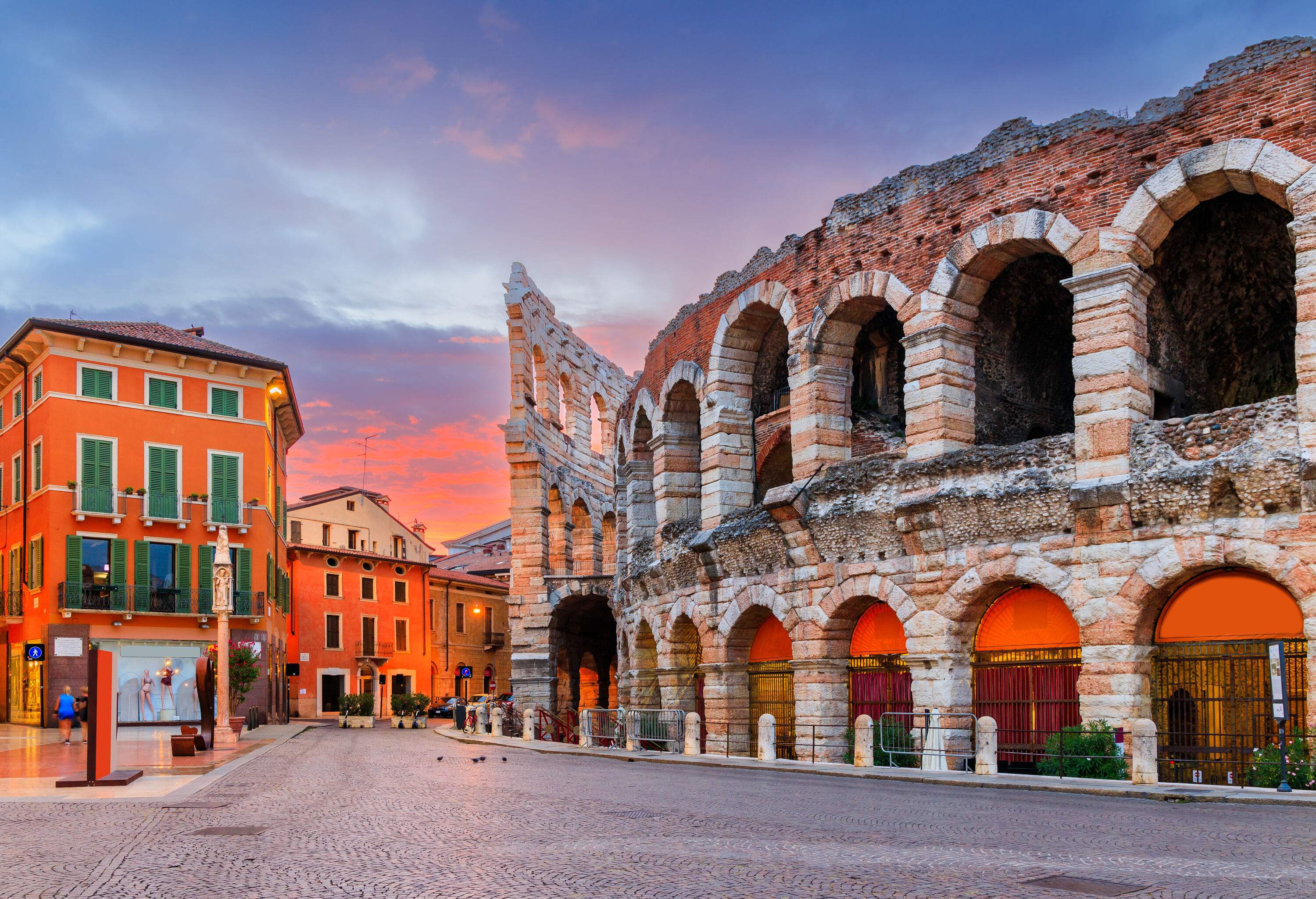 A cobbled street lined with classic buildings and an arched and curved facade of a Roman amphitheatre.
