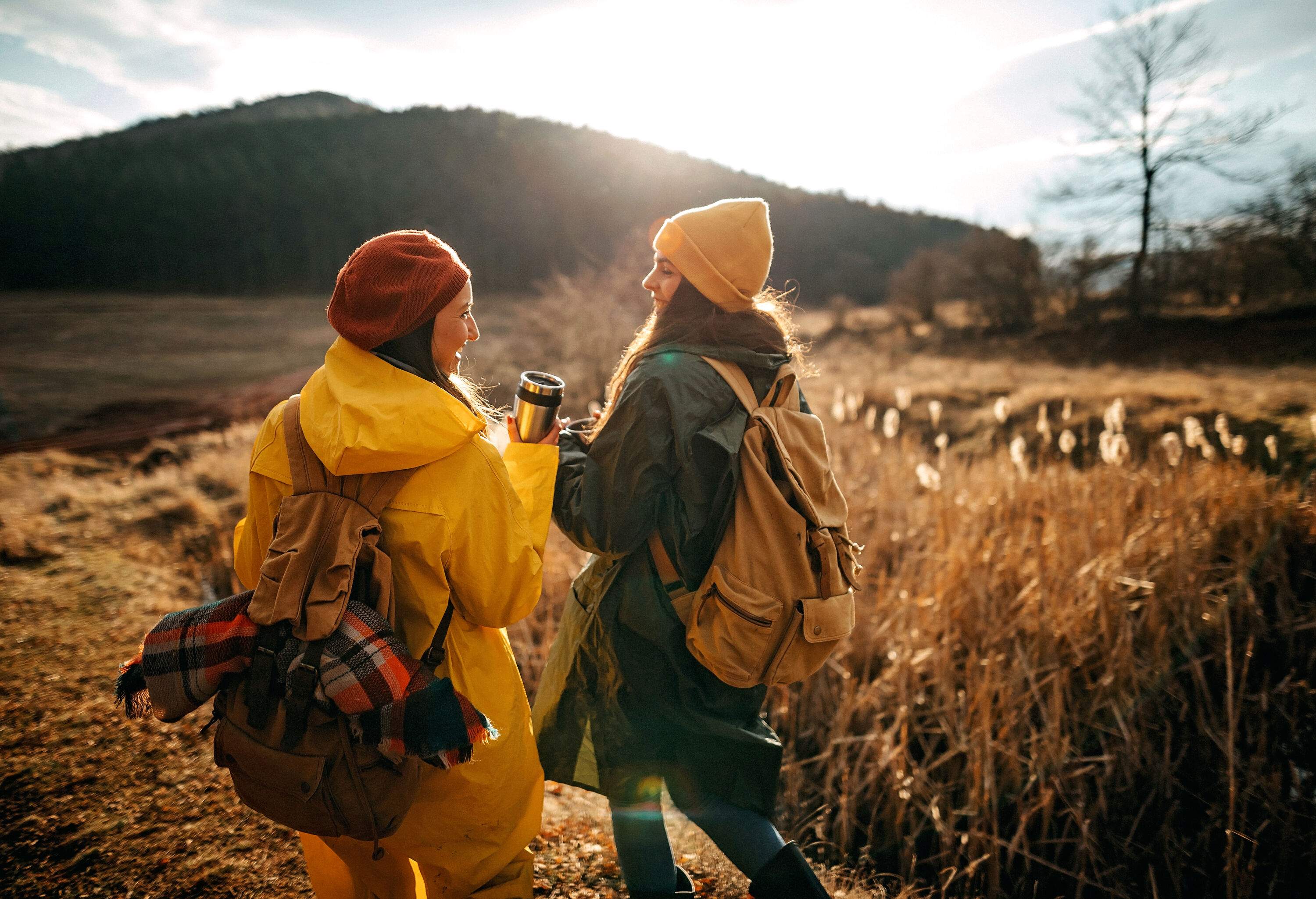 Two women on a hike wearing yellow and green raincoats, smiling at each other while holding an insulated tumbler.