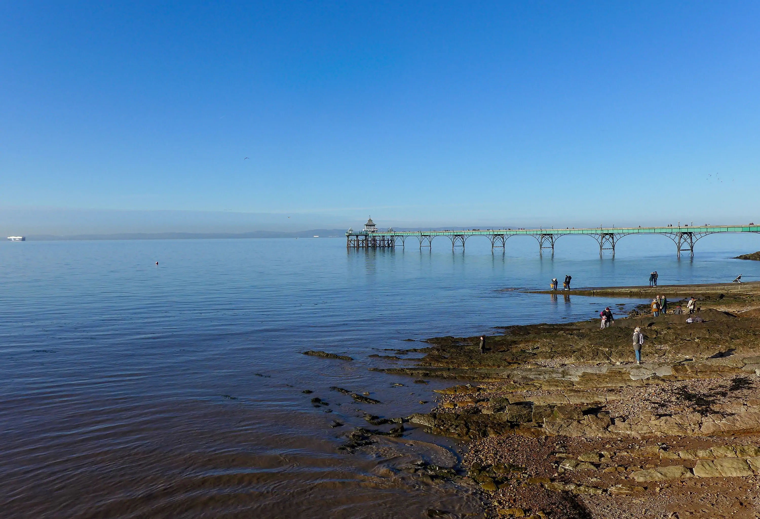People on the rocky shore overlooking a historic Victorian pier over the tranquil sea.
