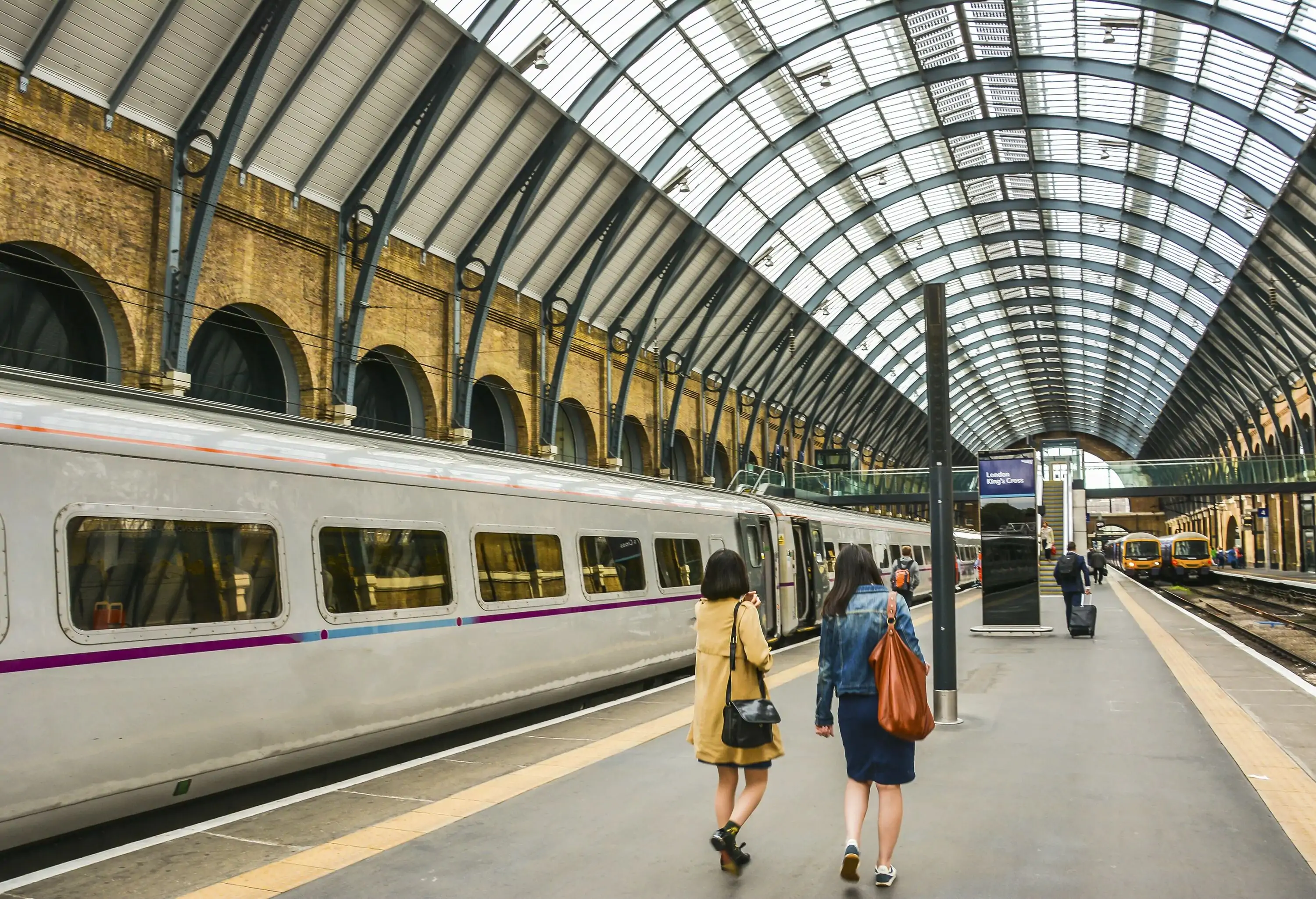 People walk on a platform beside a train under a station's brightly-lit curved ceiling.