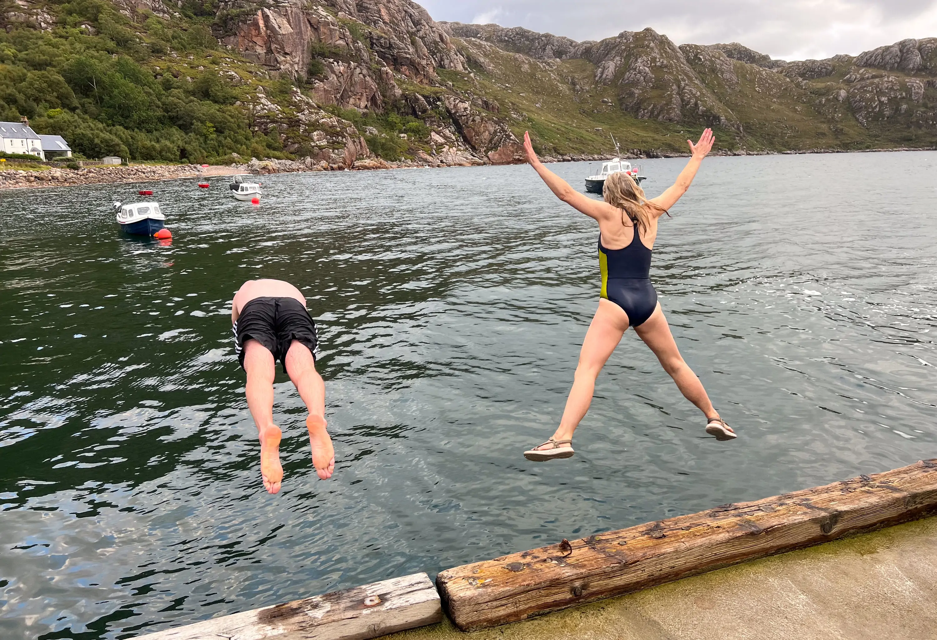 A full shot of two unrecognisable friends mid-air jumping into a loch at the same time on the side of Loch Torridon near the village of Diabaig in the west highlands of Scotland. They are wearing swimming gear.